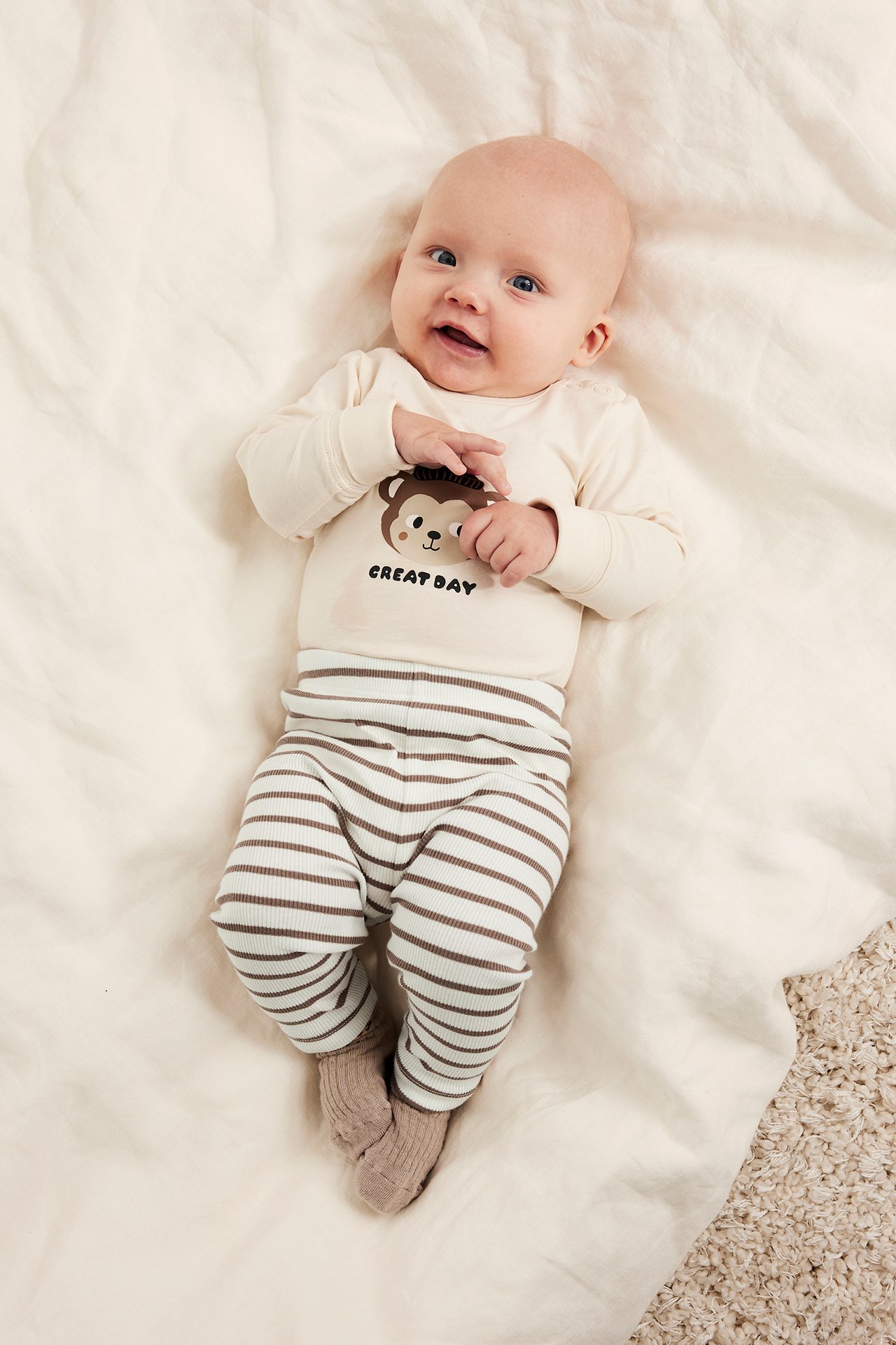 Baby bär offwhite och brunrandiga ribbade leggings, en offwhite långärmad body med apmotiv och bruna strumpor.