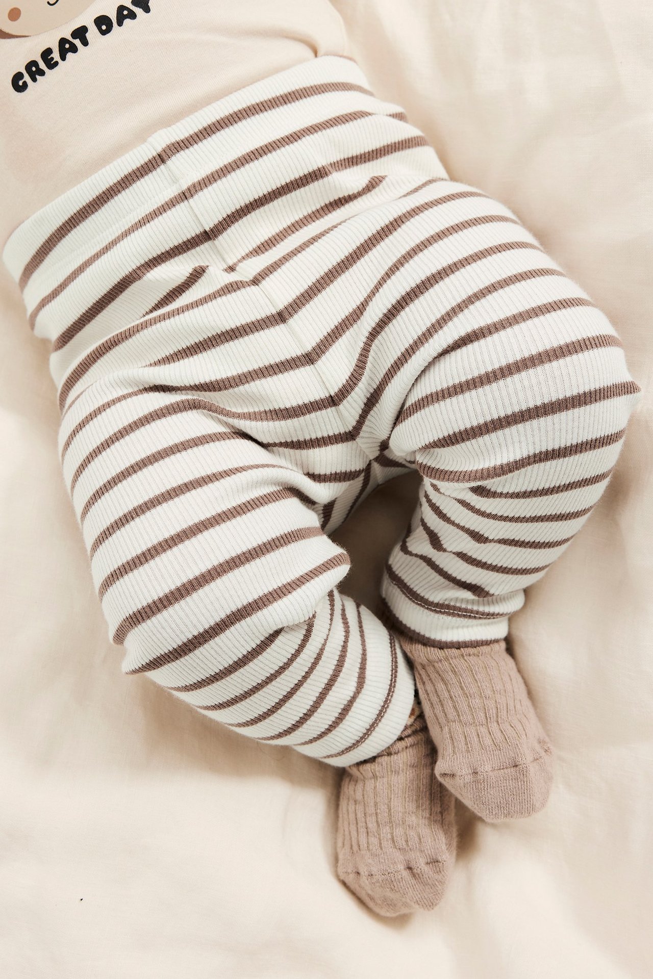Lähikuva makaavasta vauvasta, yllään luonnonvalkoiset ja ruskearaidalliset ribbileggingsit, vaalea toppi ja ribbisukat.