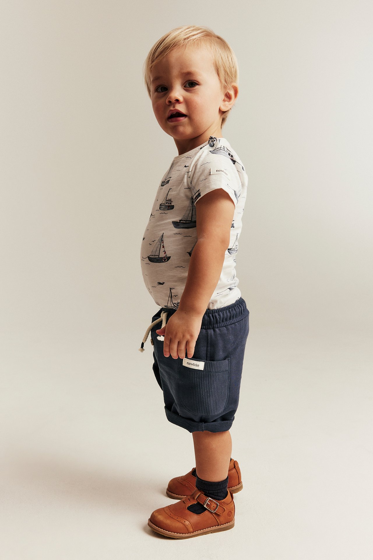 Sivukuva: vauva pukeutuu tummansinisiin slub-shortseihin, joissa resoritaskut, käännetyt lahkeet, venekuvioinen t-paita ja ruskeat kengät.