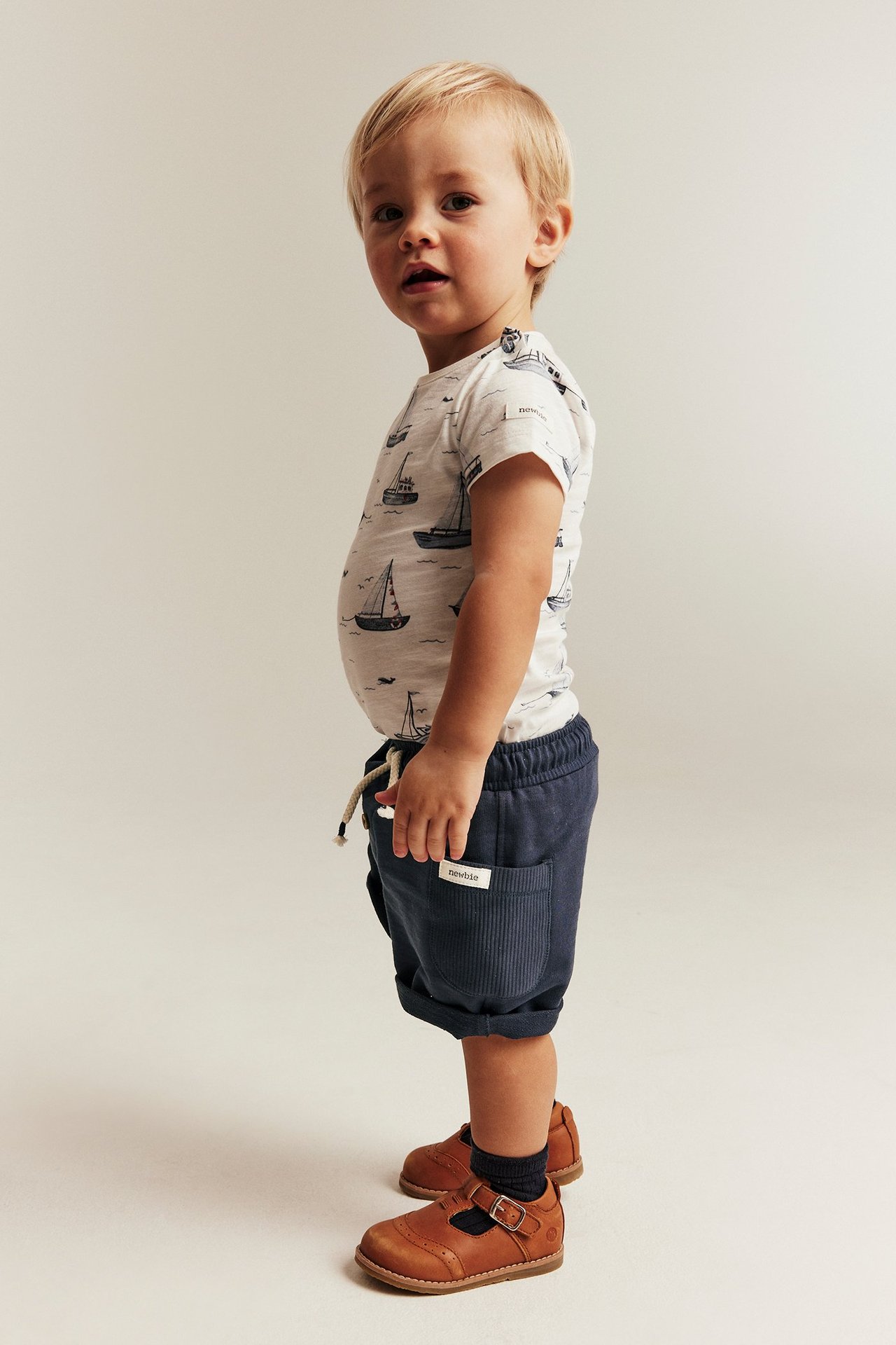 Sideutsikt: baby har på seg mørkeblå slub-shorts med ribbestrikkede lommer, oppbrettede kanter, båtmønstret t-skjorte og brune sko.