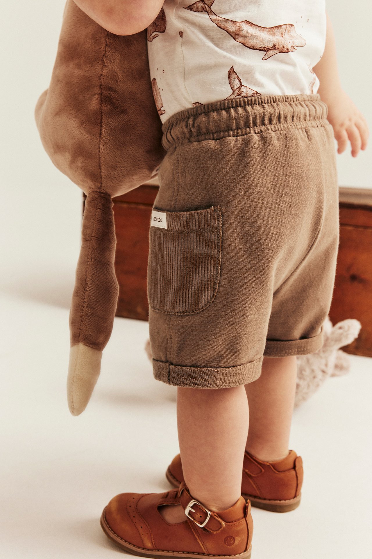 Baby i brune slub-bomullsshorts, hvalmønstret t-skjorte og brune skinnsko.