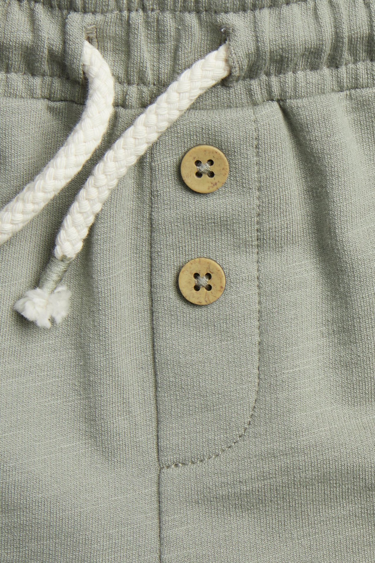 Närbild på mörkgröna shorts i slubtrikå, resår, vit dragsko och dekorativa bruna knappar.