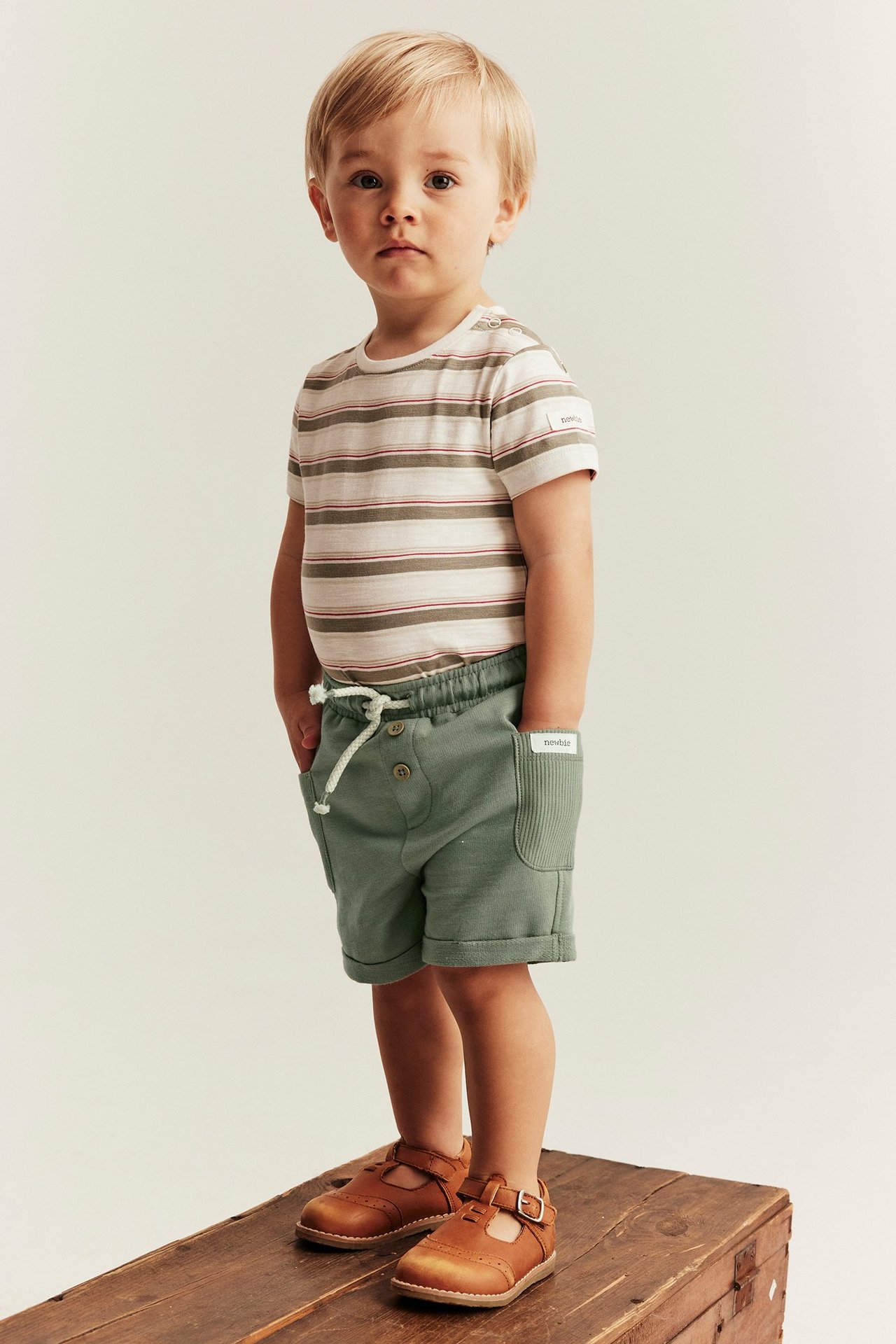 Framifrån, ett barn i mörkgröna slubtrikåshorts, en randig t-shirt och bruna T-bar skor.