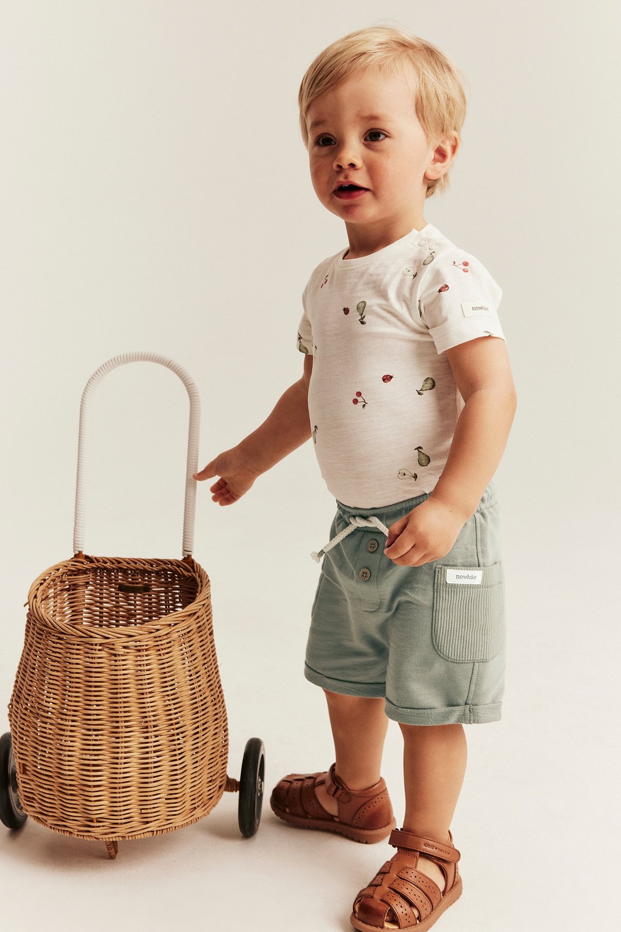 Z przodu: niemowlę w zielonych szortach slub, białej koszulce we wzory i brązowych sandałach.