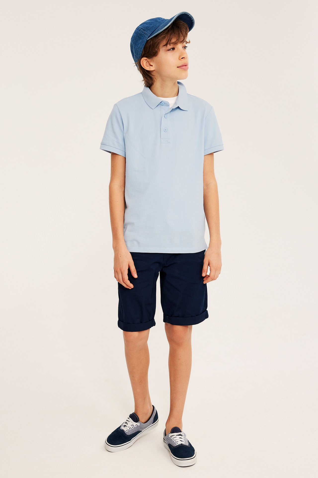 Frontbilde av et barn i lyseblå bomullspiké med knapper, marineblå shorts, denimcaps og joggesko.