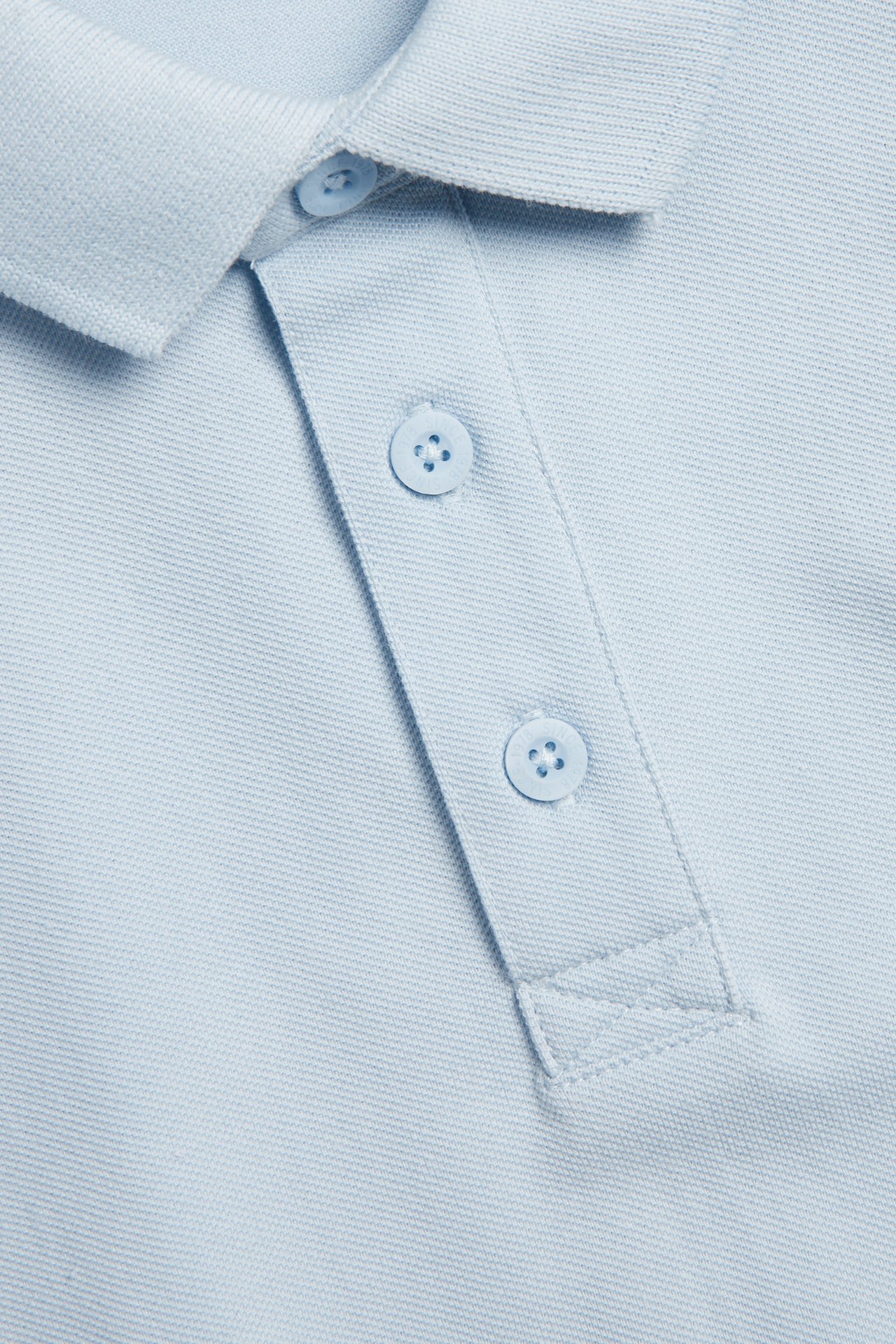 Nærbilde av en lyseblå kortermet pikétrøye i bomull med krage og knappelukking med tre knapper.