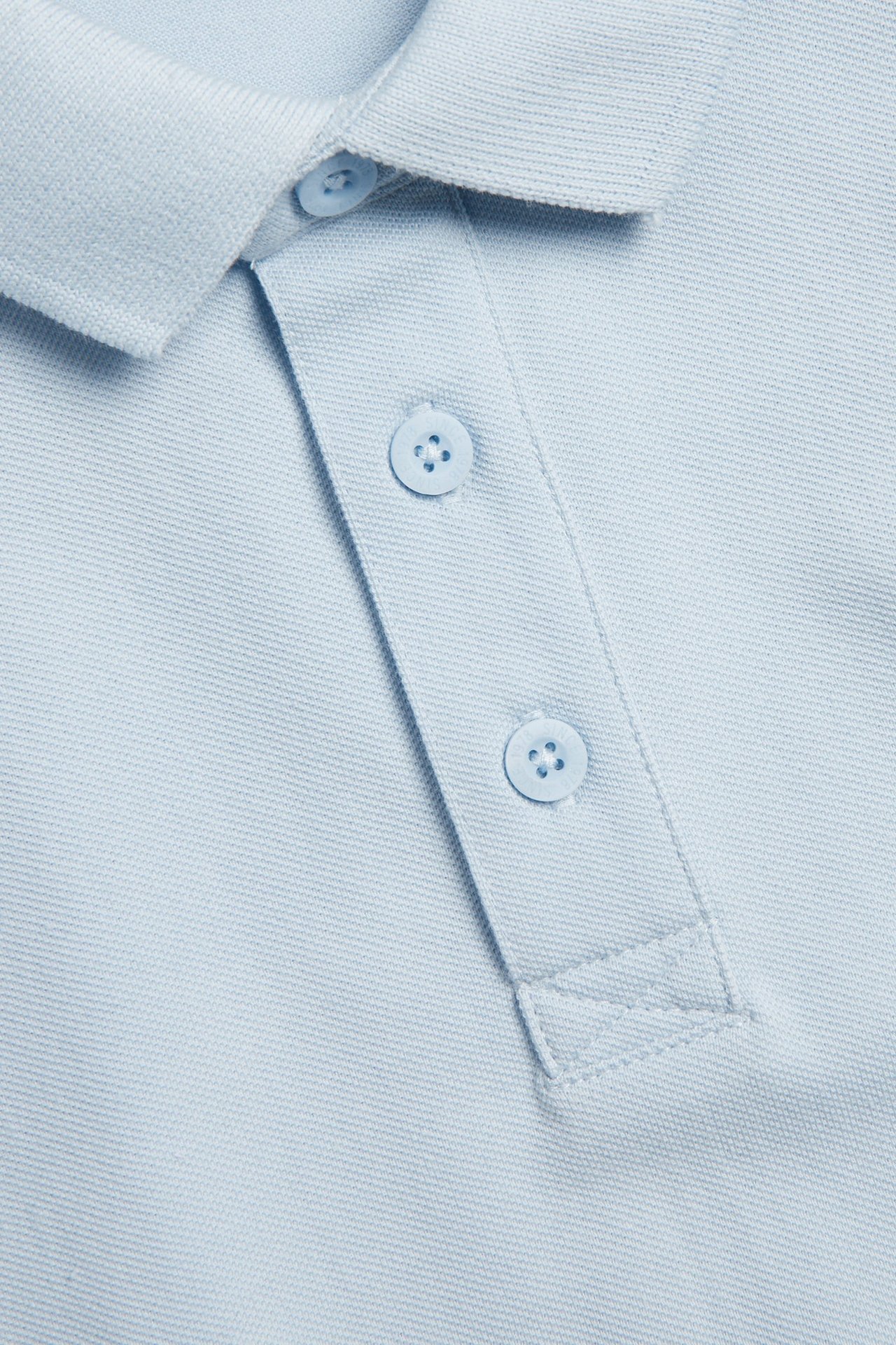Närbild av en ljusblå kortärmad pikétröja i bomull med krage och knappslå med tre knappar.