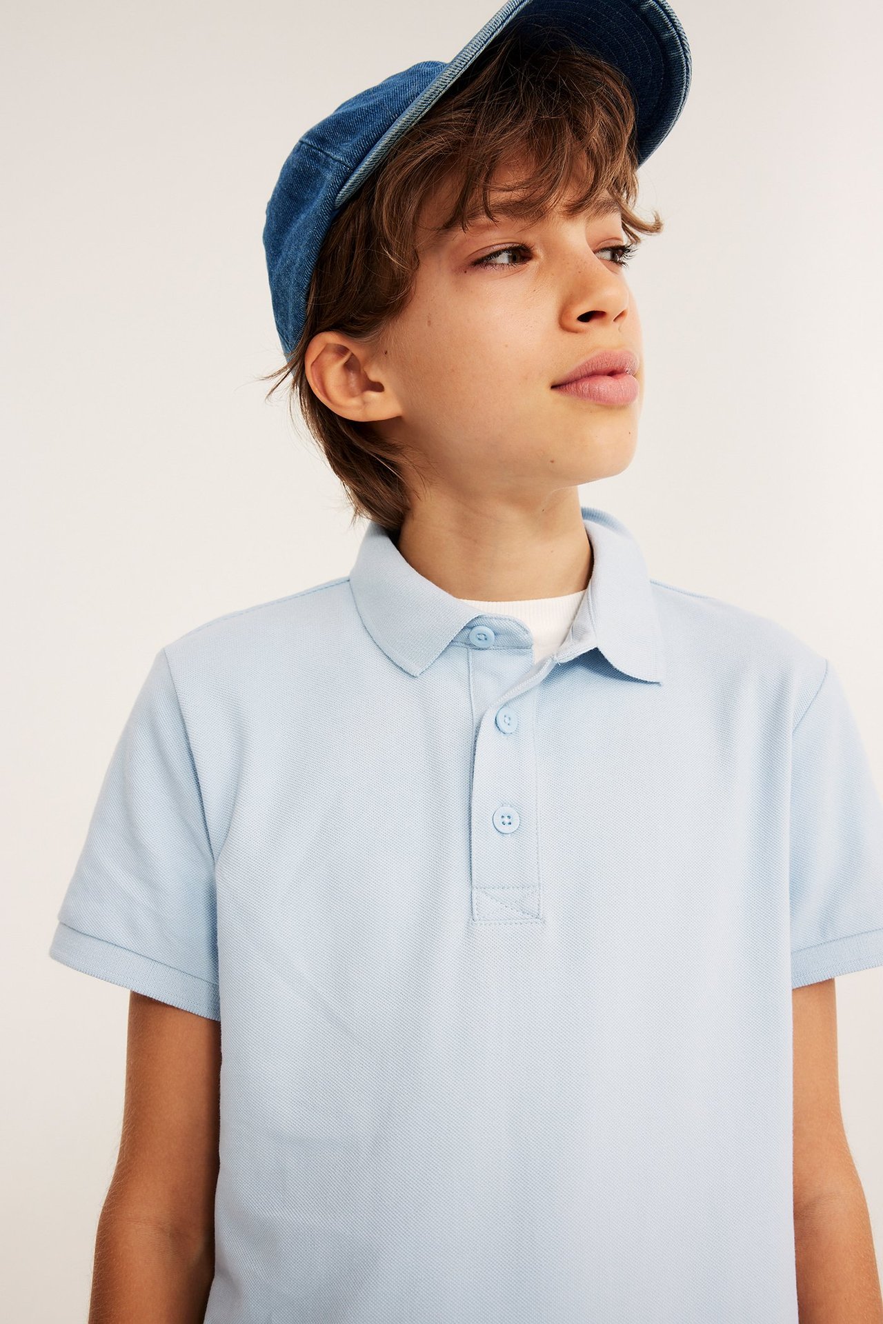 Nærbilde: Barn i lyseblå kortermet pikéskjorte i bomull med knapper og blå denimcaps.