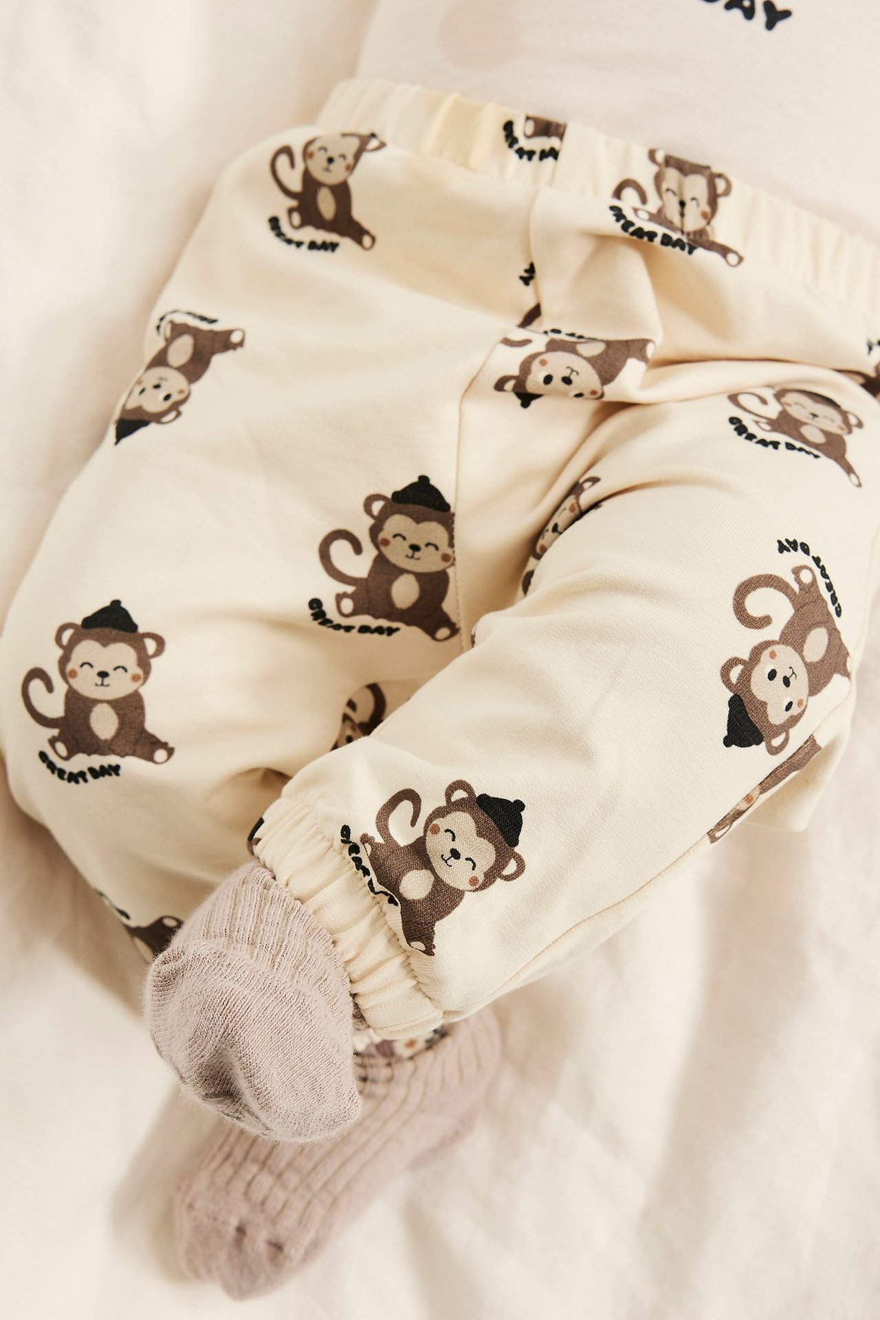 Närbild på babys ben i ljusbeige mjukisbyxor med apmotiv och ljuslila strumpor.