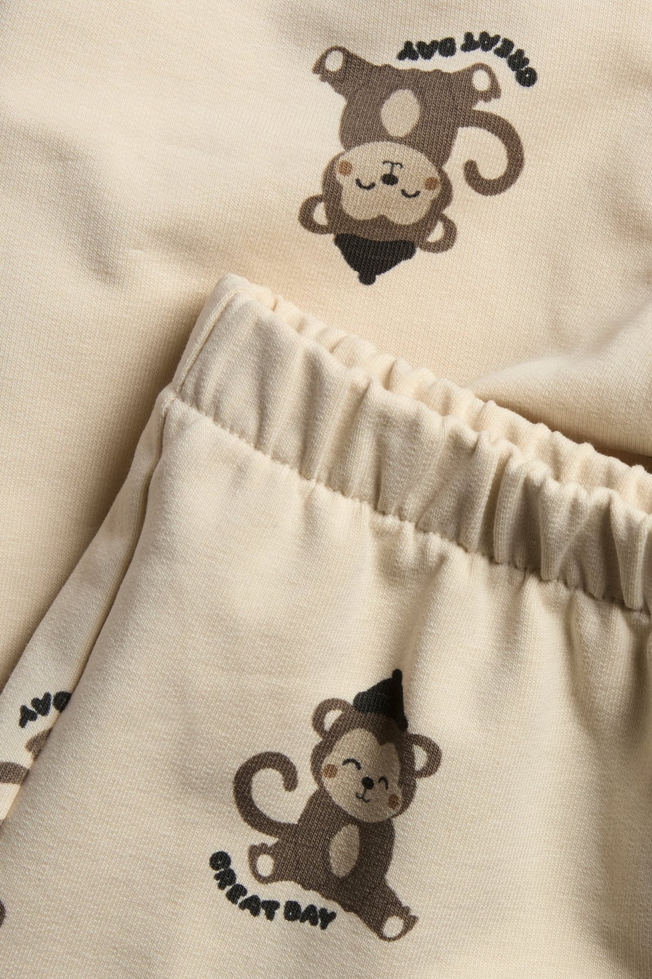 Zbliżenie na jasnobeżowe dresowe spodenki niemowlęce z brązowym nadrukiem małpek i elastycznym pasem.