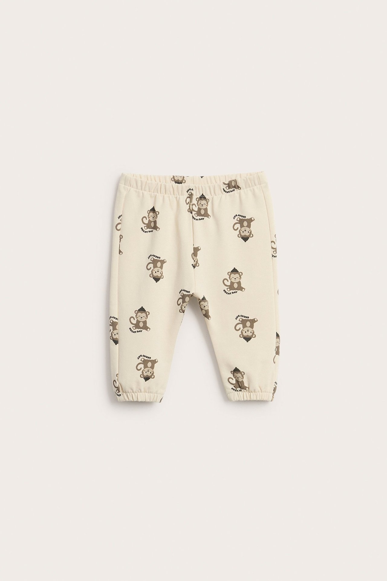 Przód jasnobeżowych spodni dresowych dla niemowląt z brązowym wzorem małpek i elastycznymi ściągaczami.
