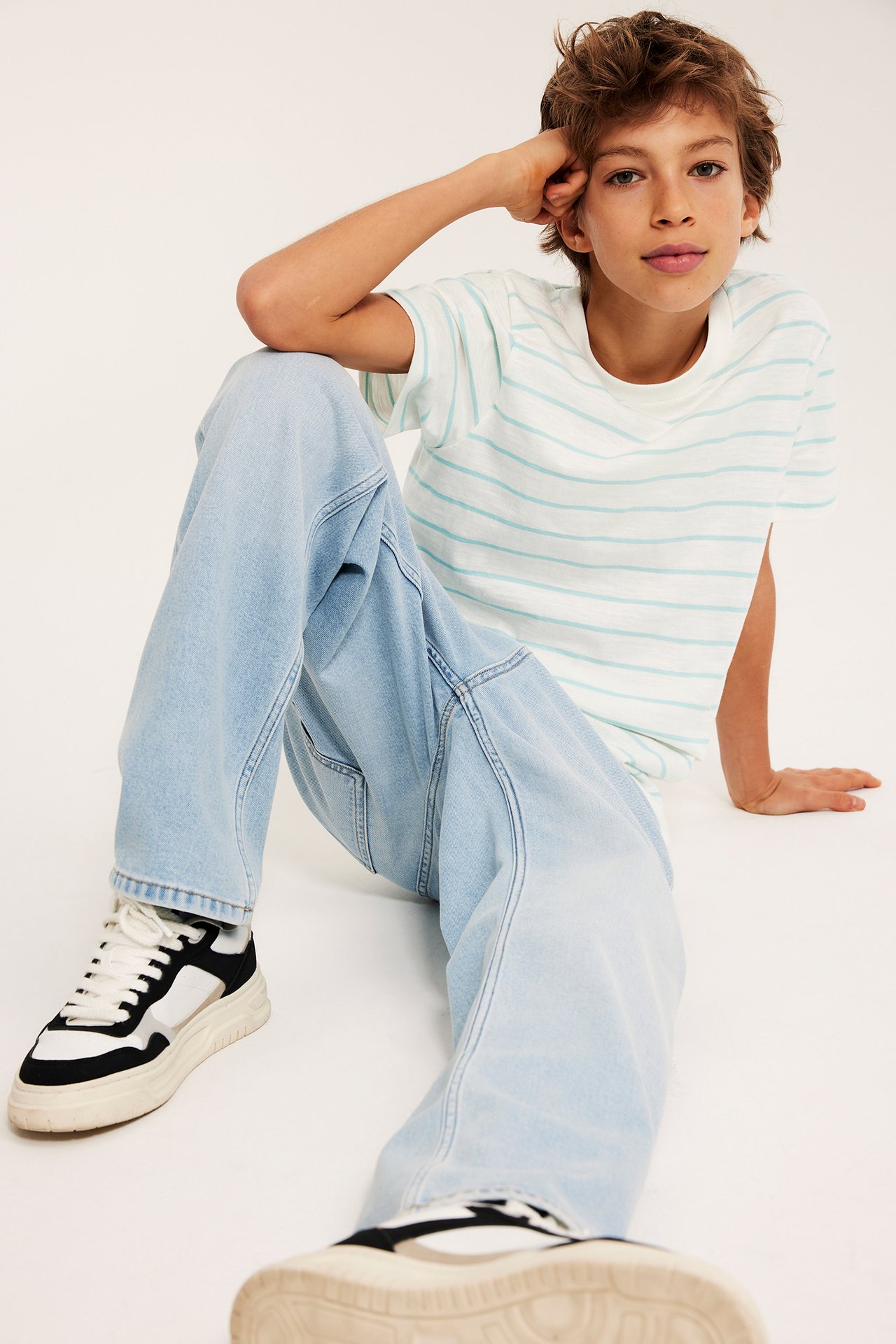 Edestä, lapsi päällä vaalea turkoosi-valkoraidallinen puuvillainen t-paita, vaaleansiniset farkut ja mustavalkoiset tennarit.