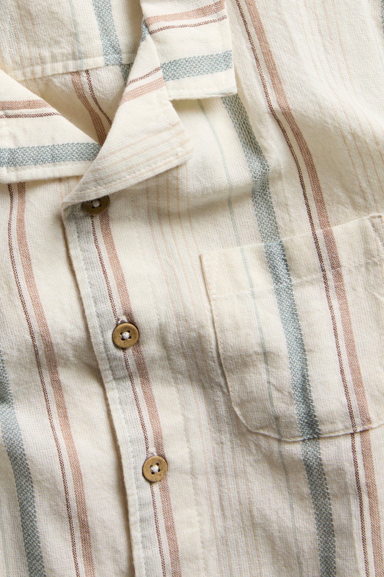 Nærbilde av offwhite stripete kortermet vevd skjorte med krage, treknapper og brystlomme.