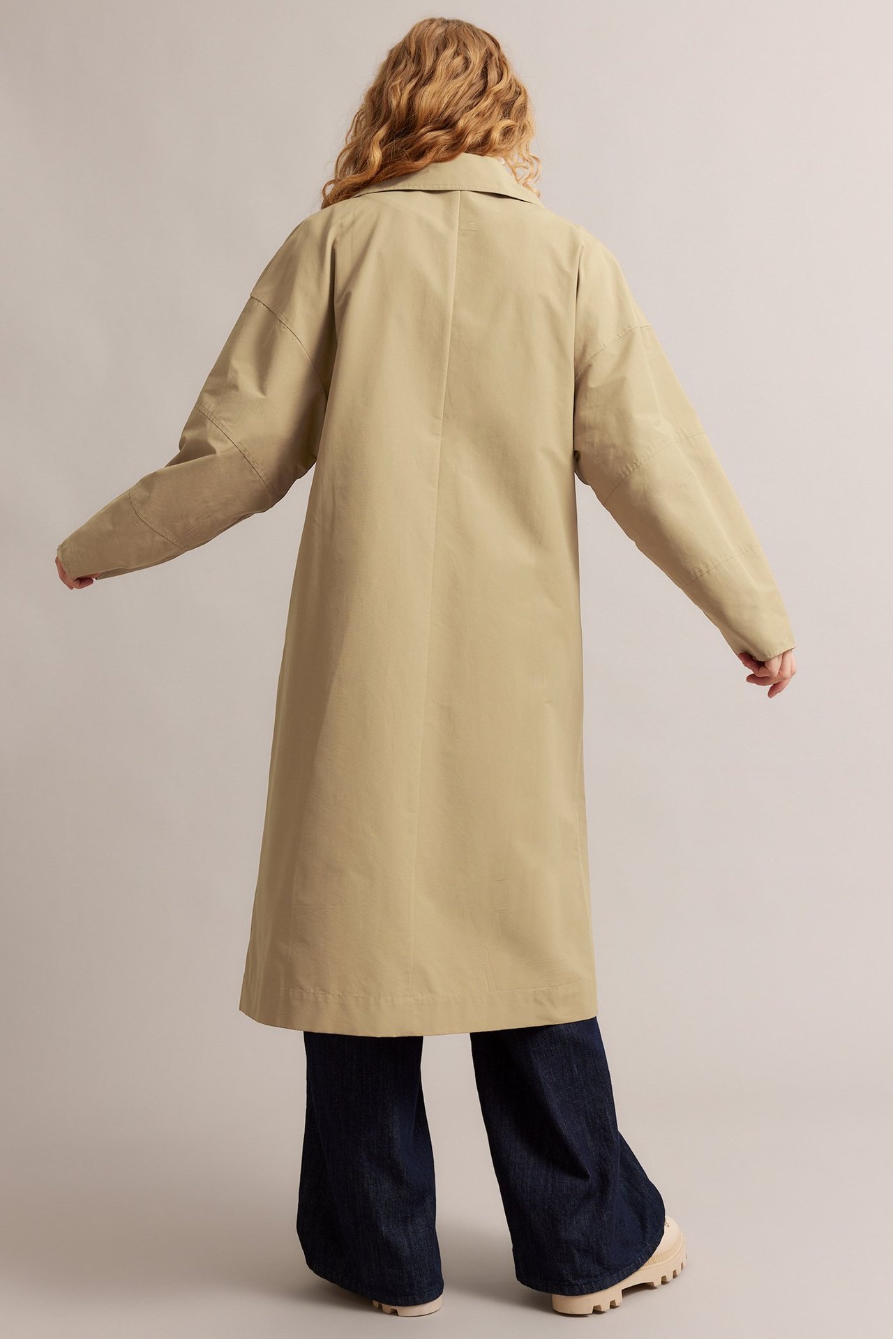 Widok z tyłu: Kobieta w beżowym trenczu z bawełny, ciemnoniebieskich dżinsach i jasnobeżowych butach.