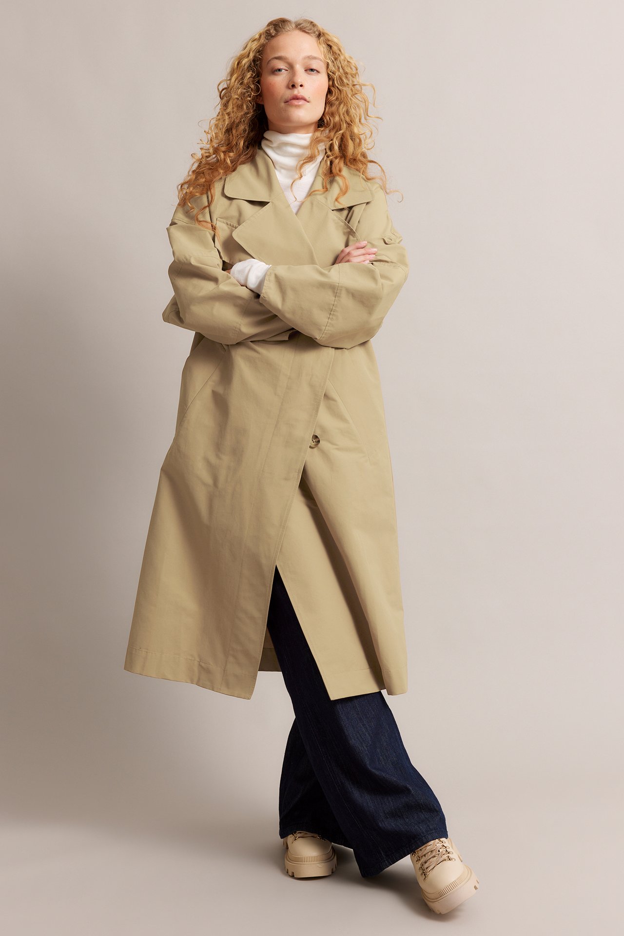 Forfra: Kvinne i beige bomullstrenchcoat over hvit høyhalset genser, mørke jeans og beige sko.
