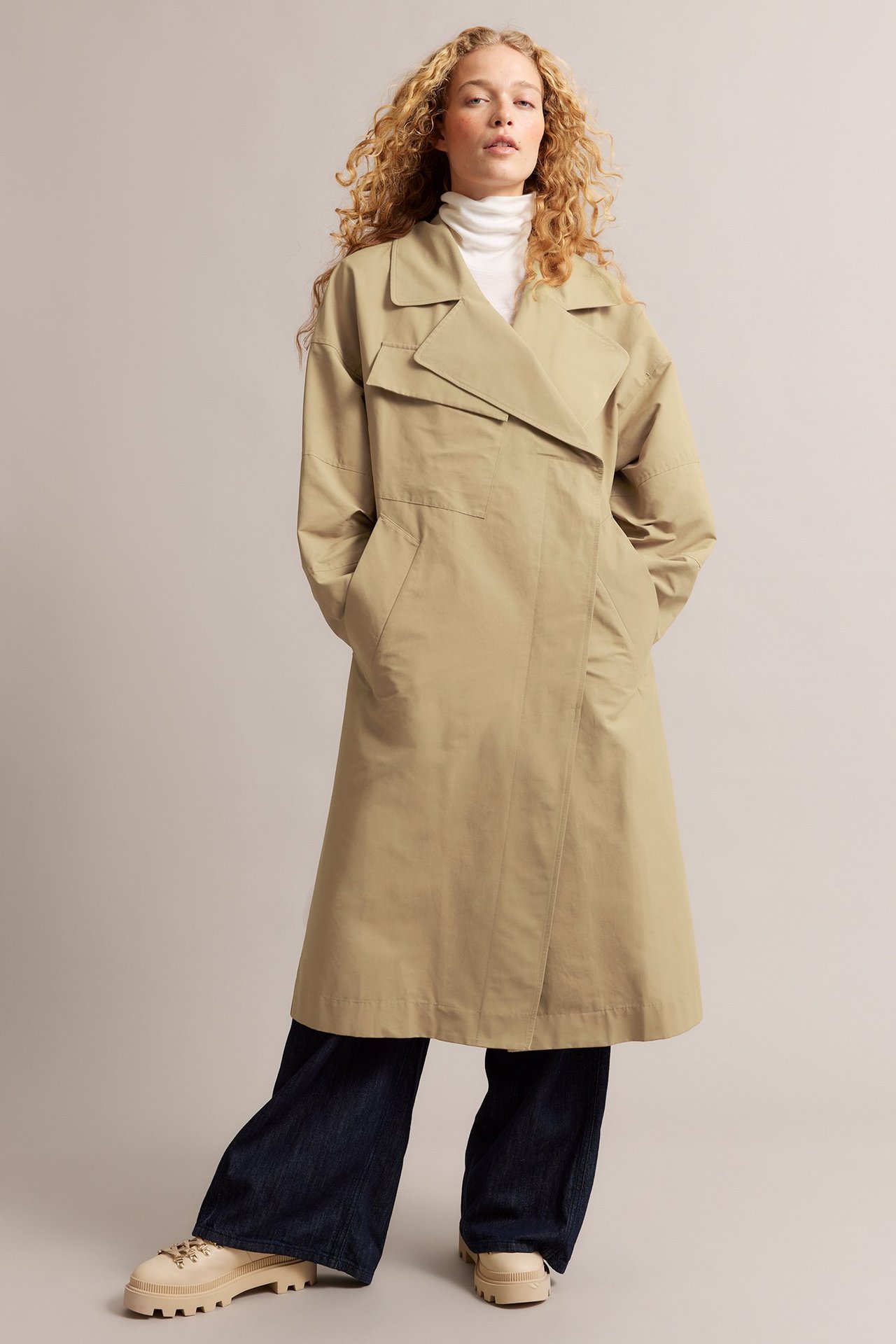 Widok z przodu: Kobieta w luźnym beżowym trenczu, białym golfie, ciemnych szerokich jeansach i masywnych beżowych botkach.