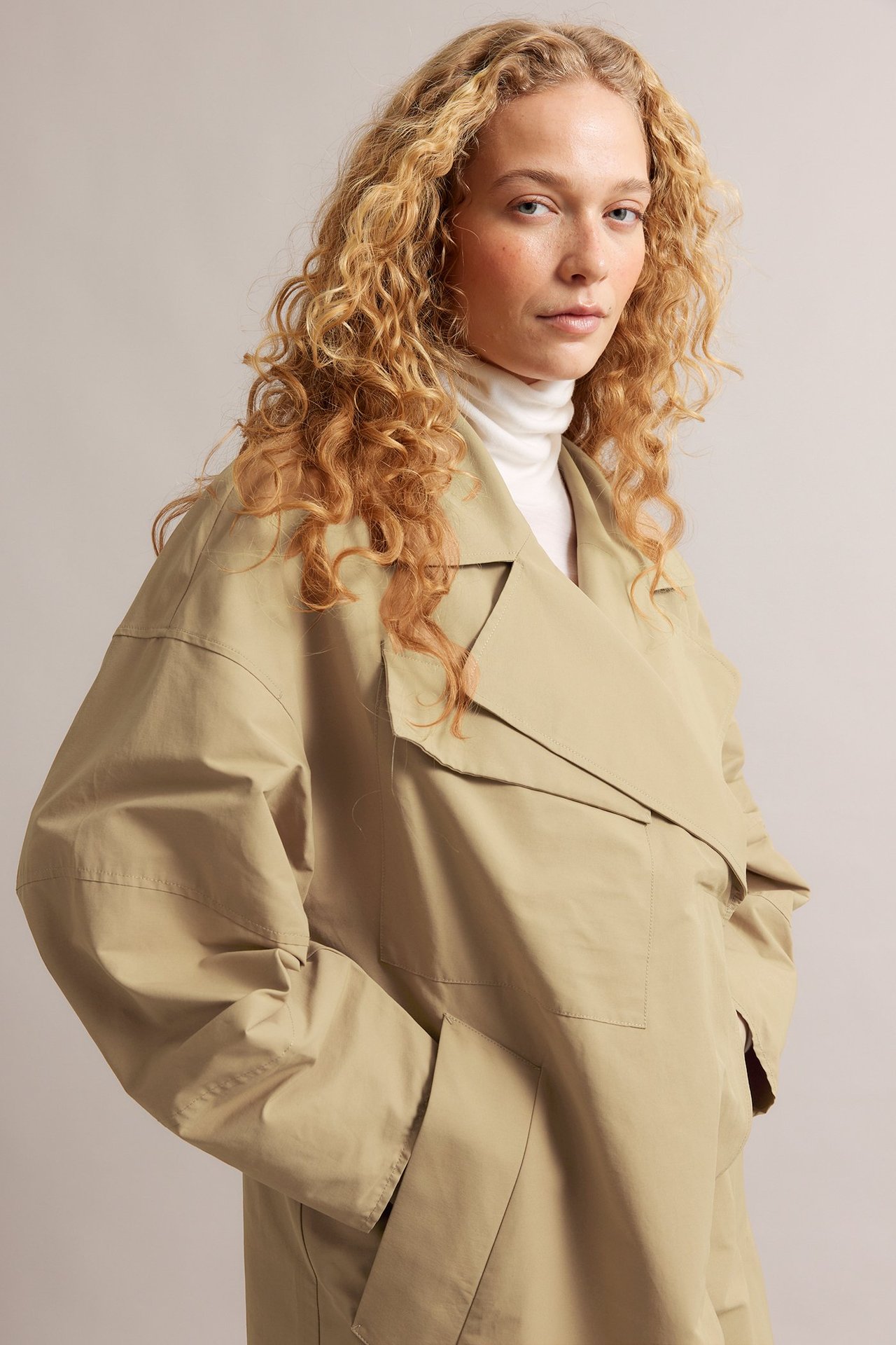 Widok z przodu: kobieta w beżowym trenczu z mieszanki bawełny z przednimi kieszeniami na białym golfie.