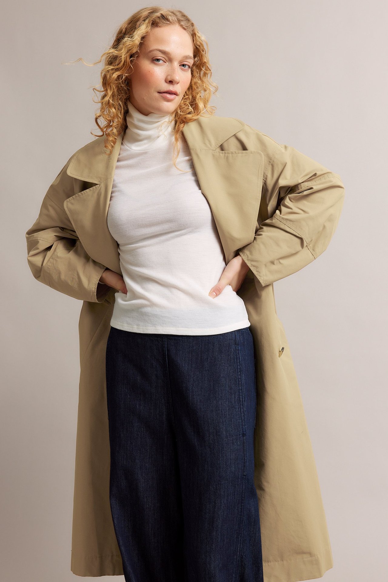 Widok z przodu: kobieta w otwartym beżowym trenczu z mieszanki bawełny, na białym golfie i ciemnoniebieskich spodniach.