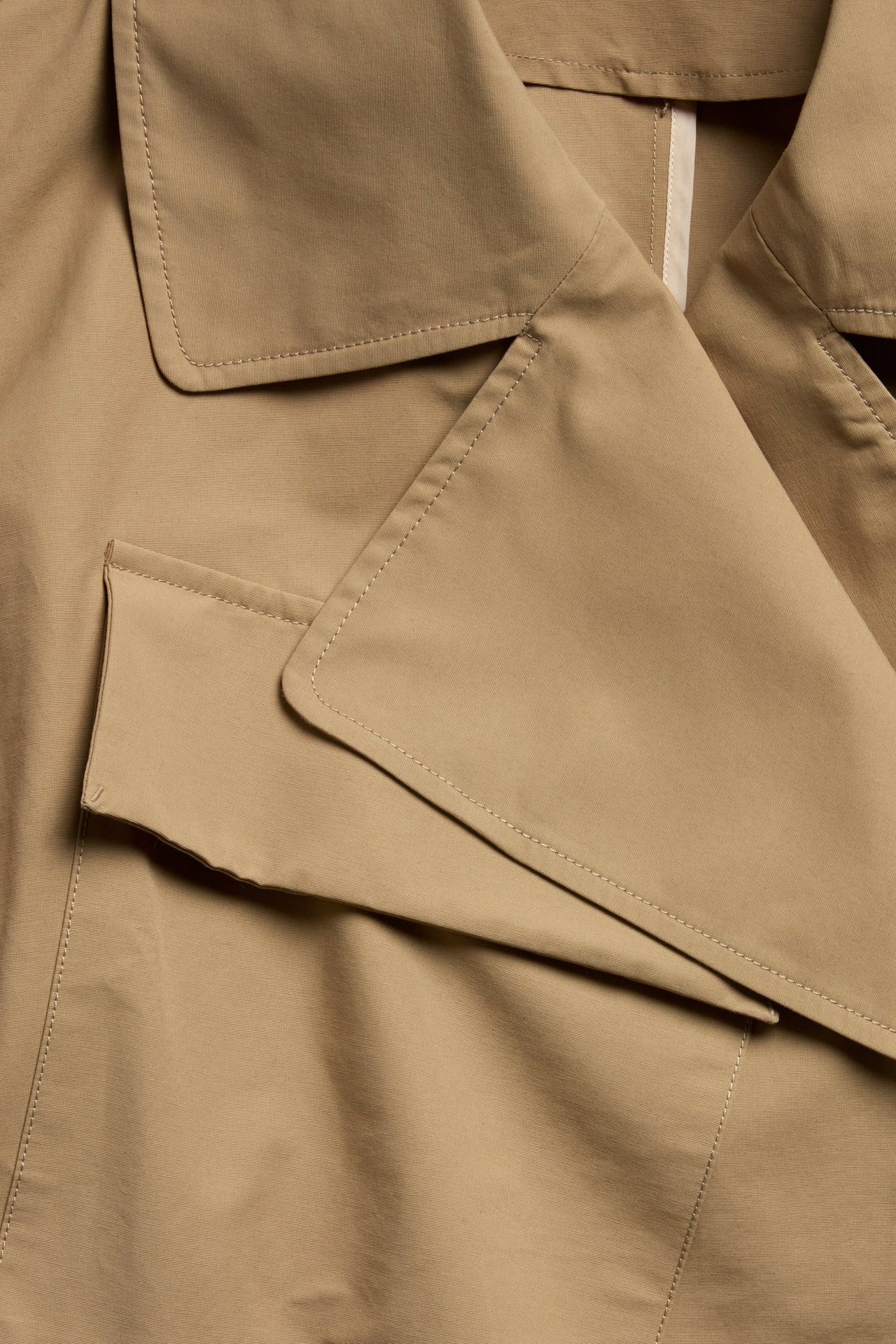 Nærbilde av en beige trenchcoat i bomullsblanding med bred krage og synlig lommeklaff.