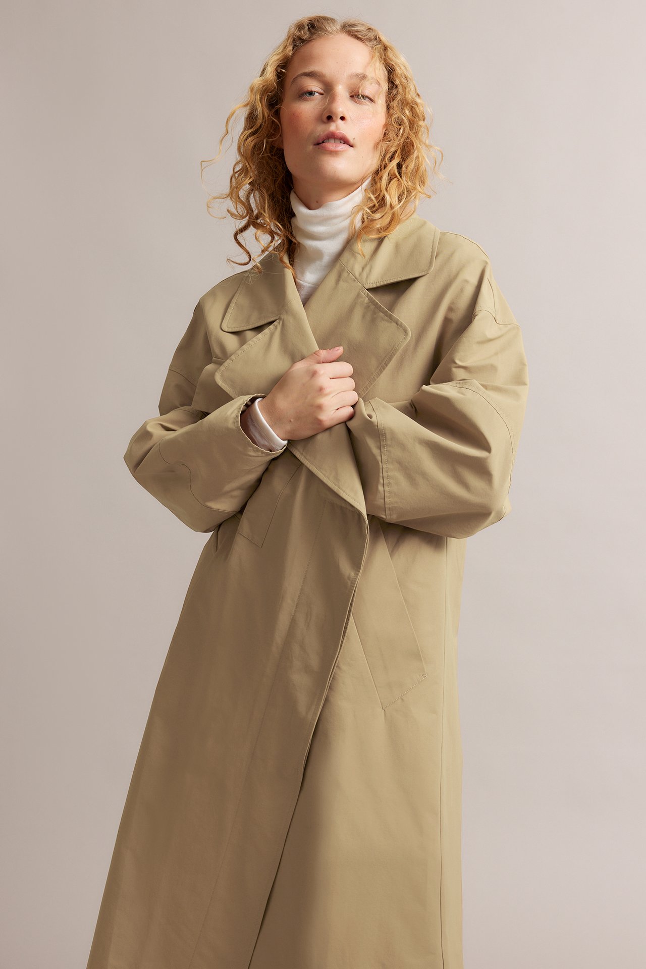 Forfra: kvinne iført beige trenchcoat i bomullsblanding over en hvit høyhalset genser, holder den lukket.
