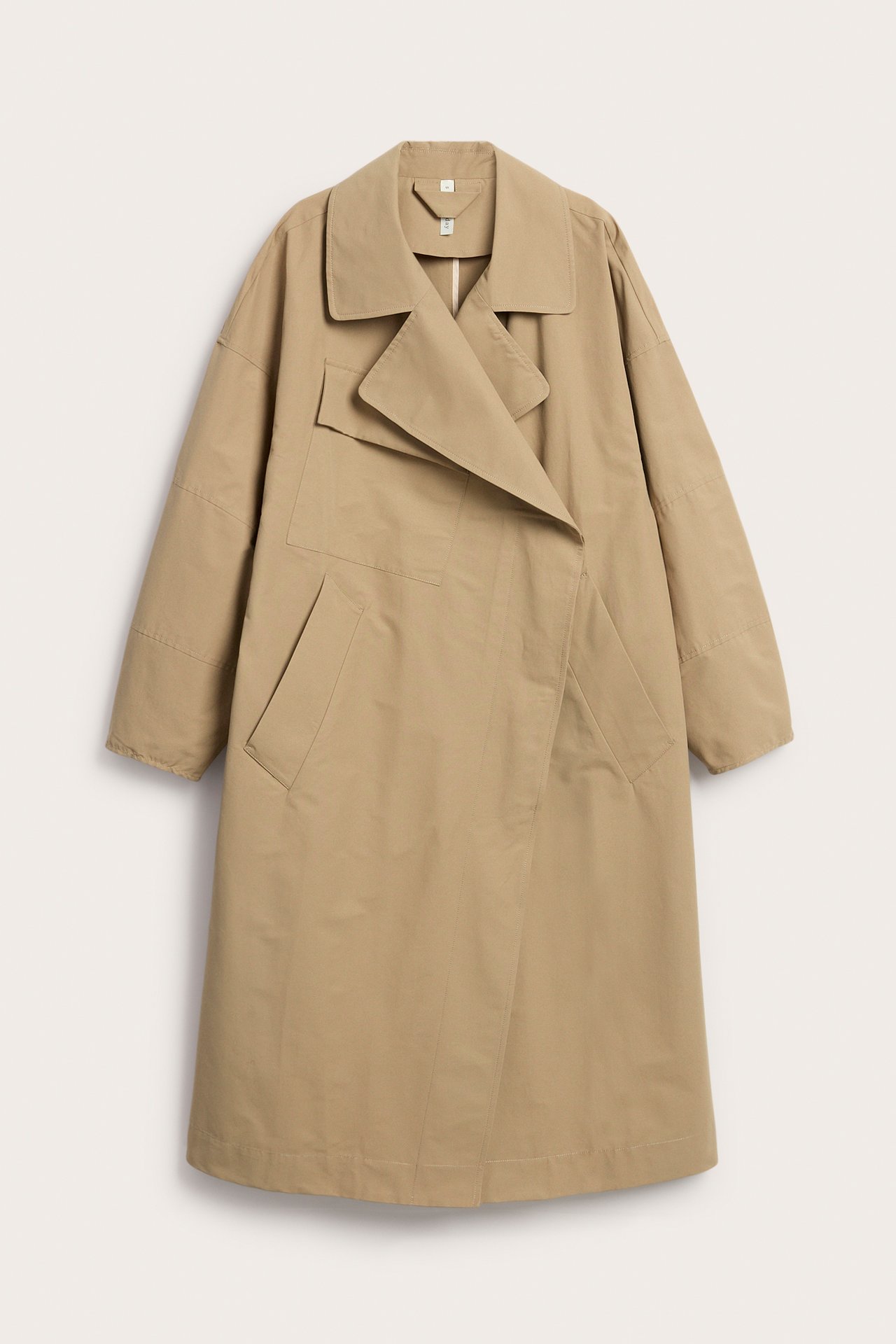 Framifrån: Beige trenchcoat i bomullsblandning med asymmetrisk front, breda slag och fickor fram.