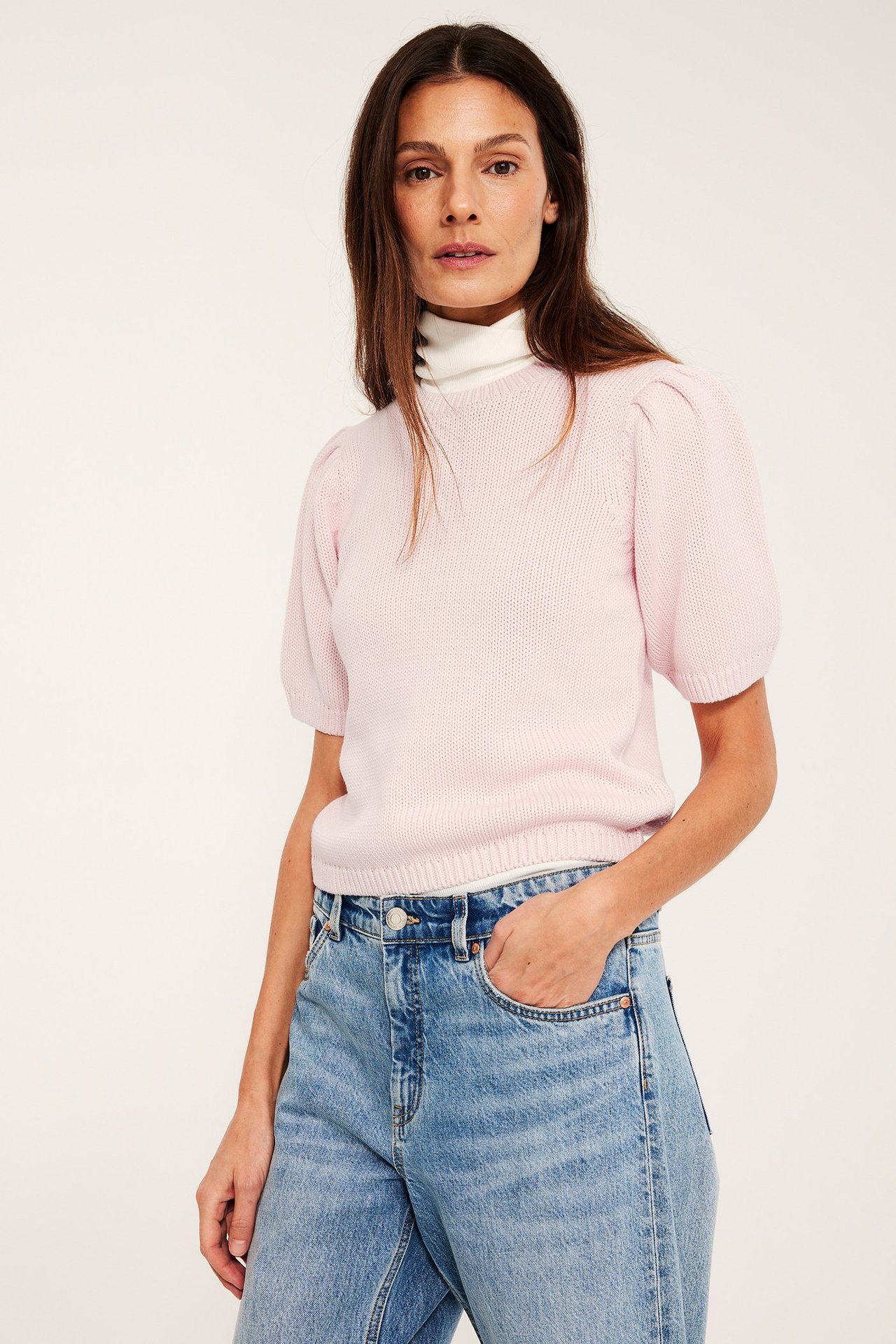 Frontbilde av kvinne i lysrosa bomullsstrikket genser med puffermer, over en hvit turtleneck og blå jeans.