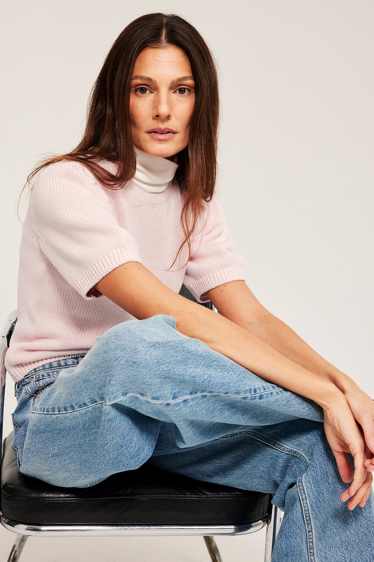 Etunäkymä: Nainen yllään vaaleanpunainen puuvillaneulepusero, valkoisen korkeakauluksen päällä, sekä vaaleansiniset farkut.