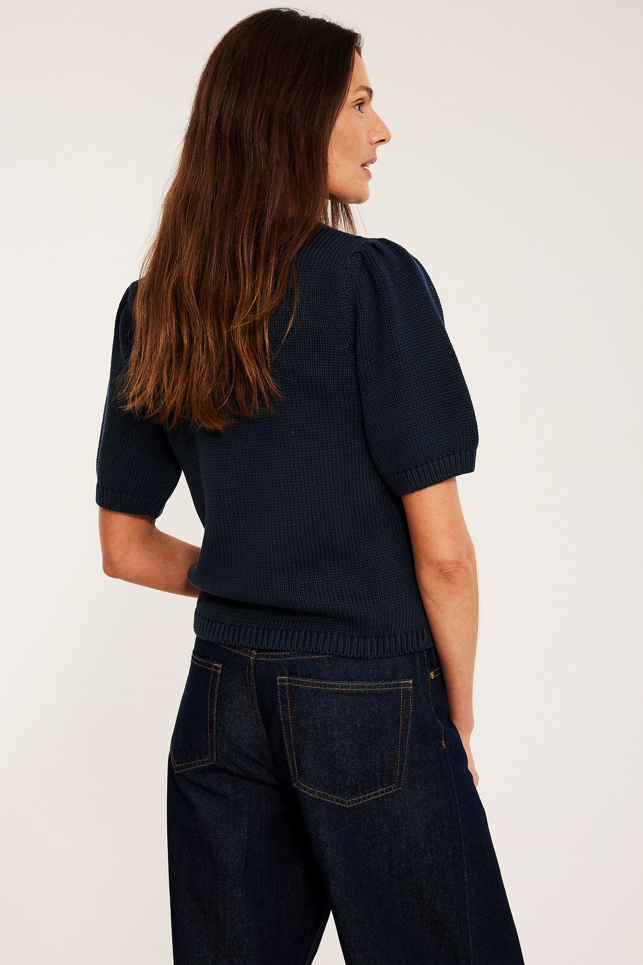 Nainen takaa, tummansinisessä puuvillaneuleessa, jossa lyhyet pöyhkeät hihat, ja tummansinisissä farkuissa.