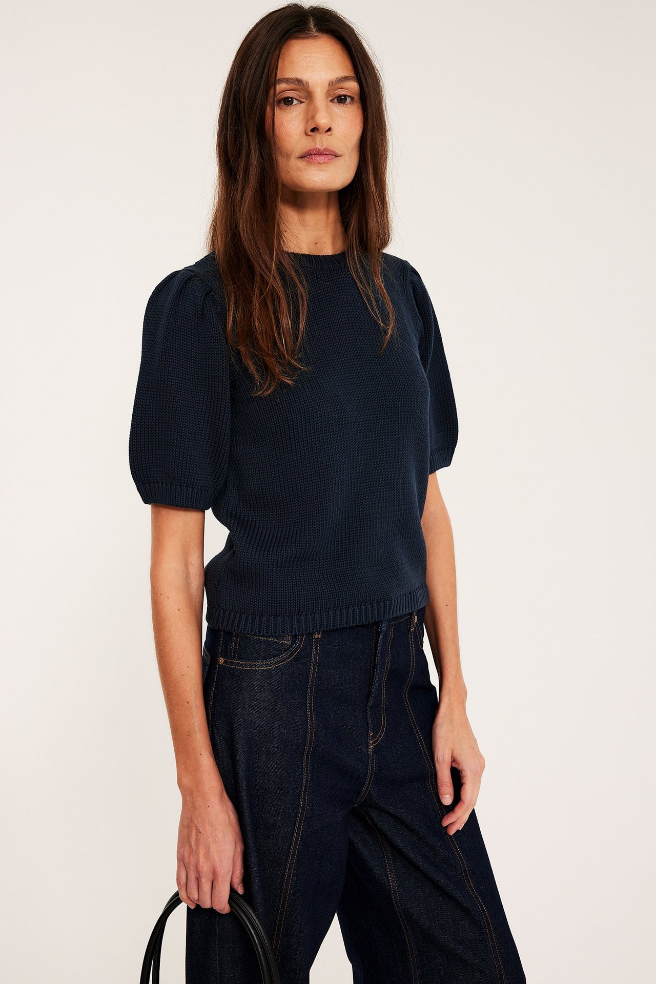 Forfra: kvinne i mørkeblå bomulls strikkegenser med korte puffermer og mørkeblå jeans.