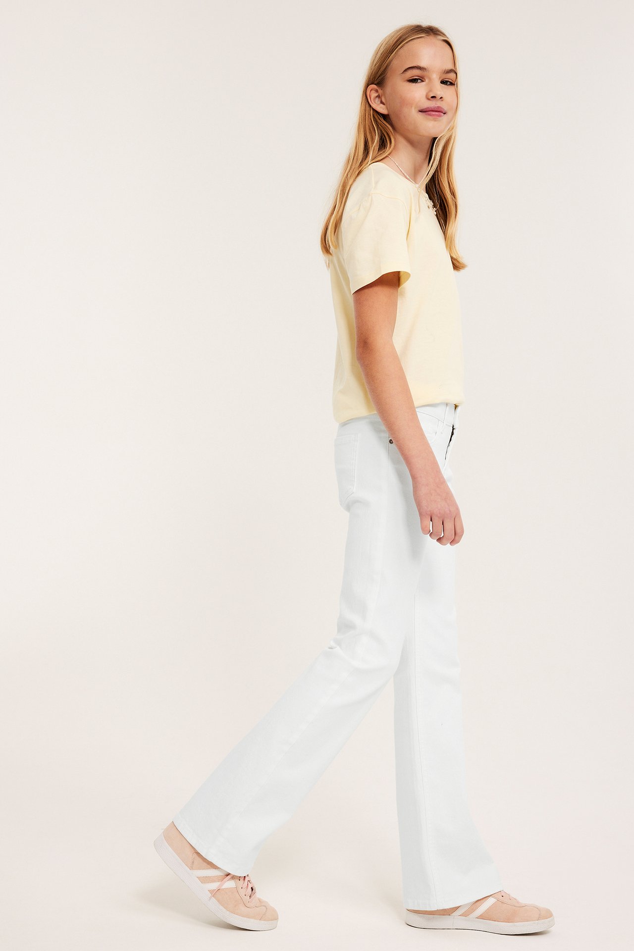 Sidevisning av et barn i hvite bootcut-jeans, lysegul t-skjorte og rosa joggesko.