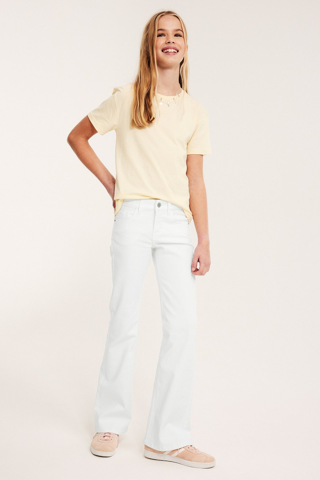 Frontvy: Barn i vita bootcut lågmidjade jeans, ljusgul t-shirt och rosa sneakers.