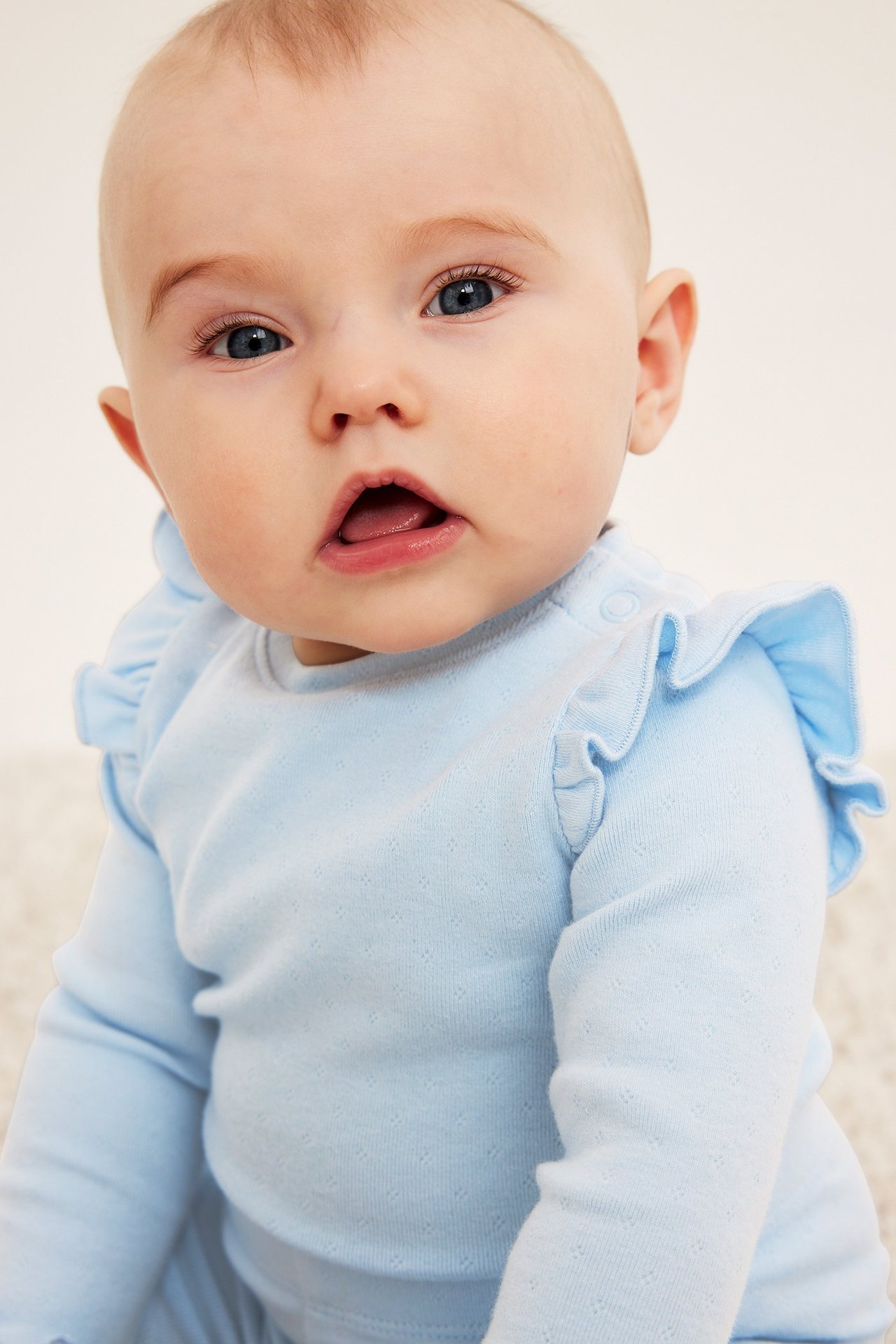Lähikuva vauvasta, yllään vaaleansininen pointelle-neuleinen pitkähihainen body olkafrilloilla.