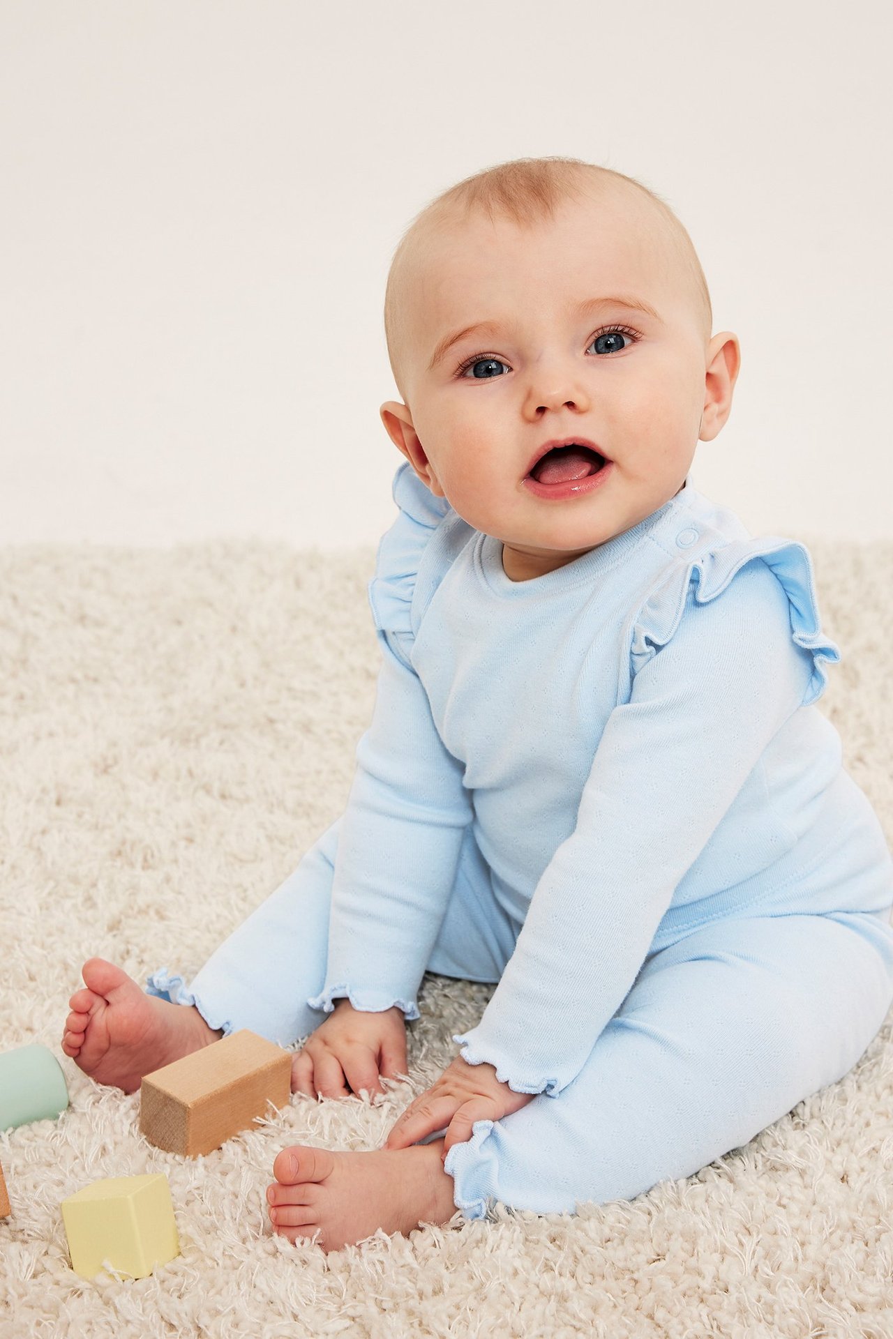 Edestä kuvattu vauva yllään vaaleansininen pitkähihainen body olkarypytyksillä ja aaltoilevilla reunoilla.