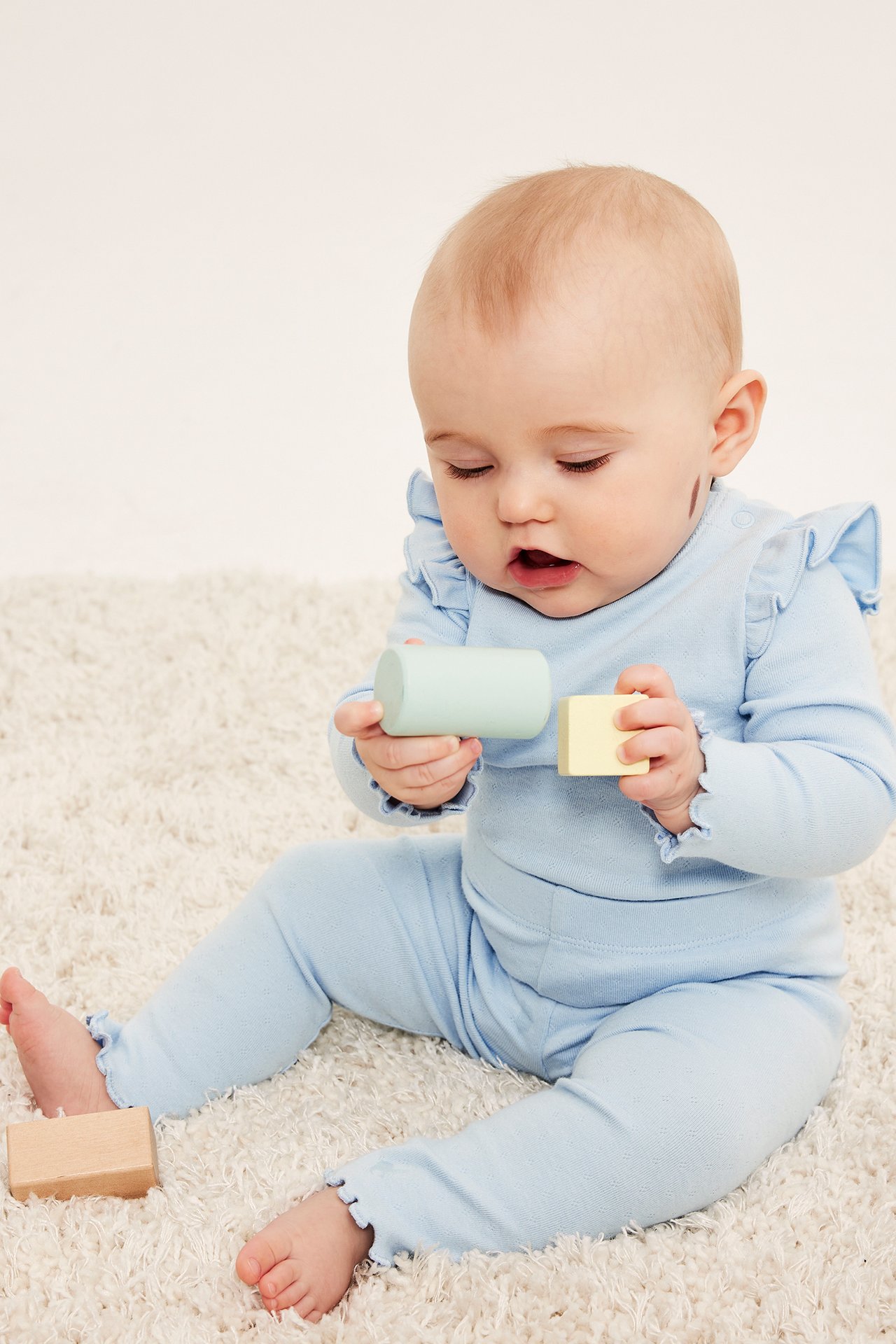 Etunäkymä, vauva yllään vaaleansiniset pointelle-leggingsit röyhelöreunoilla ja yhteensopiva toppi, pitäen palikoita.