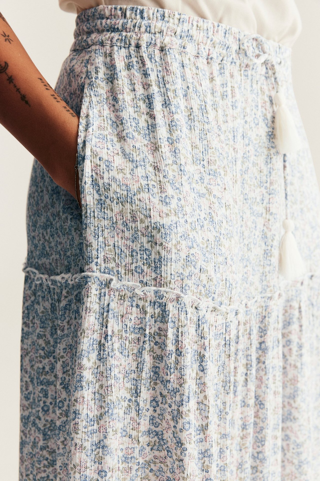 Nærbilde, sideutsikt: Kvinne i offwhite blomstret langskjørt i krøllviskose, elastisk midje, dusker og lomme.