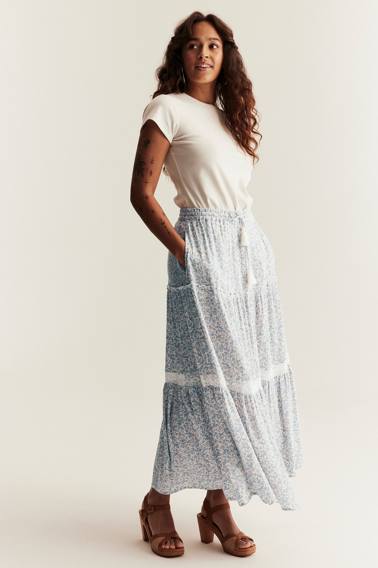 Widok z przodu: Kobieta w jasnoniebieskiej kwiecistej spódnicy maxi z wiskozy z koronką, białym t-shircie i brązowych sandałach.