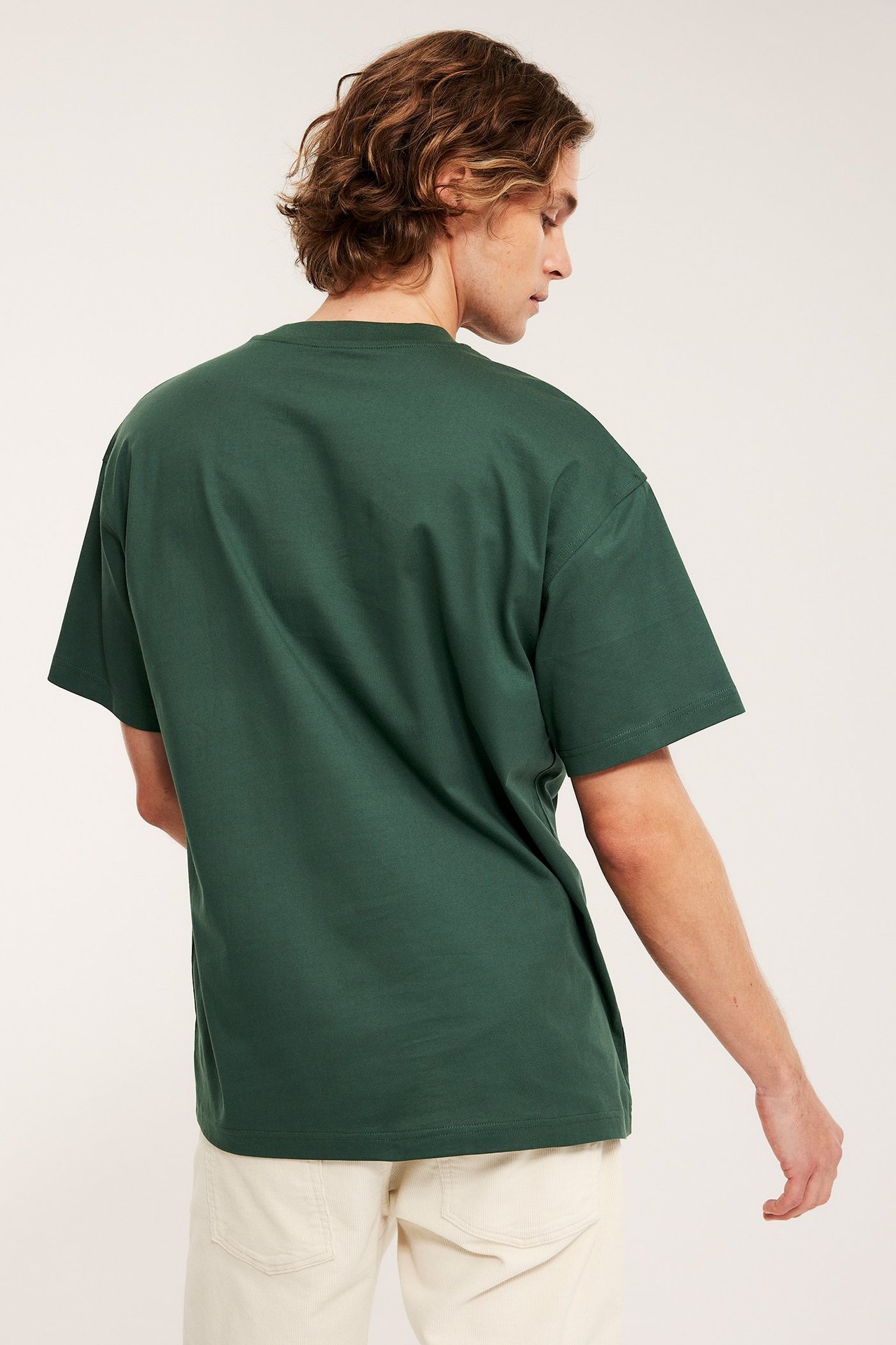Mies, takaapäin/sivulta, yllään tummanvihreä väljä puuvillainen T-paita ja vaaleanbeiget housut.