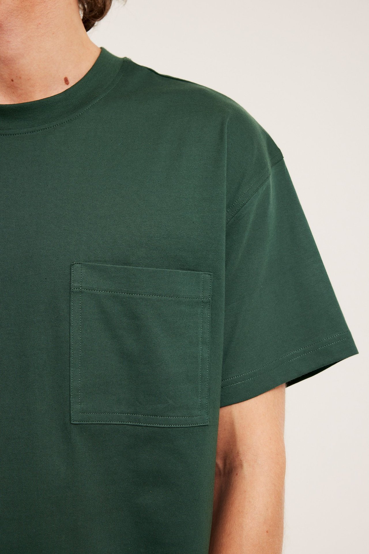 Lähikuva miehestä yllään väljä vihreä puuvillainen t-paita rintataskulla ja lyhyillä hihoilla.