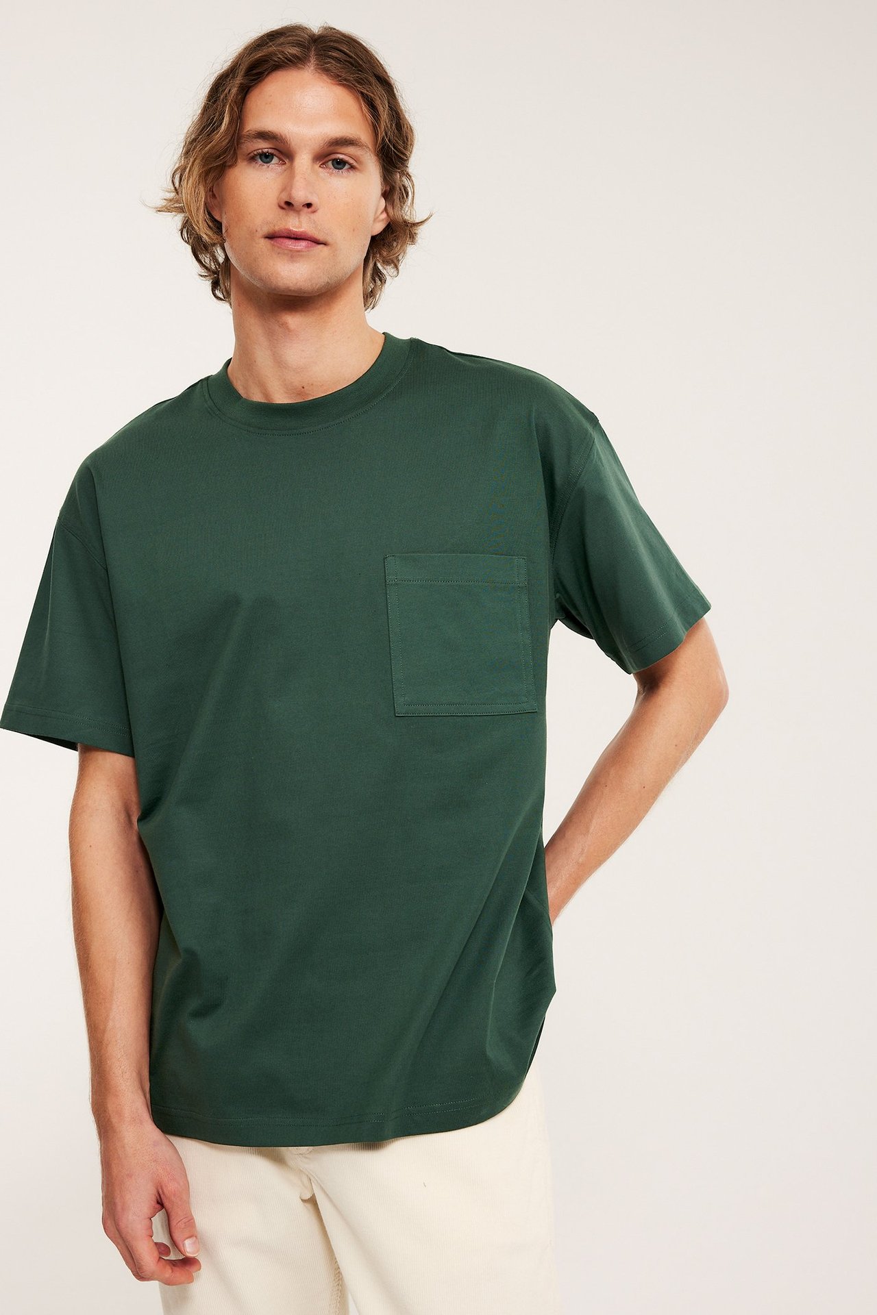 Widok z przodu mężczyzny w luźnym zielonym bawełnianym T-shircie z kieszenią, stylizowanym z jasnymi beżowymi spodniami.