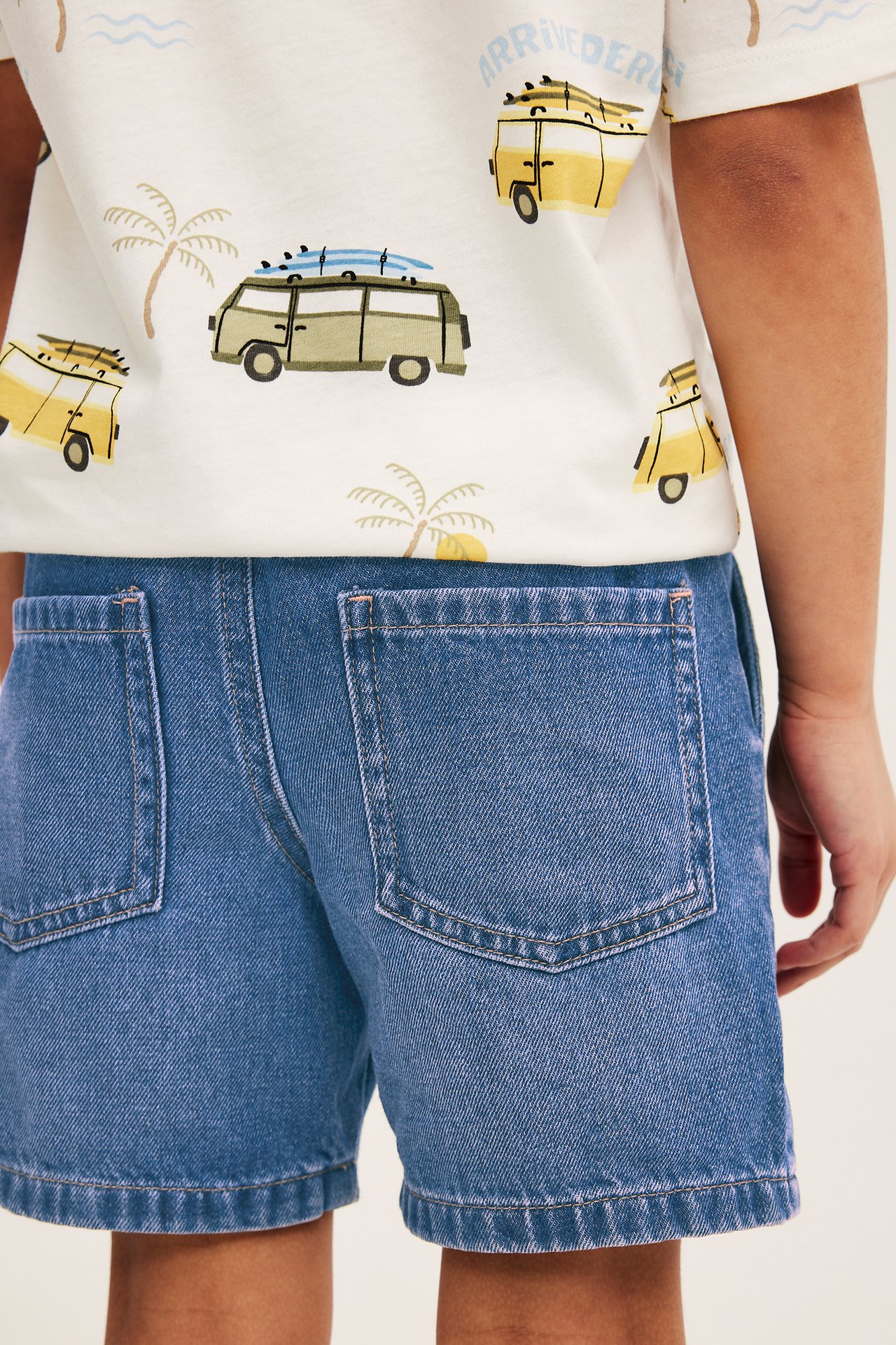 Ryggvy av ett barn i mörk denimblå dra-på-shorts med bakfickor och en vit t-shirt med surfbilsmönster.