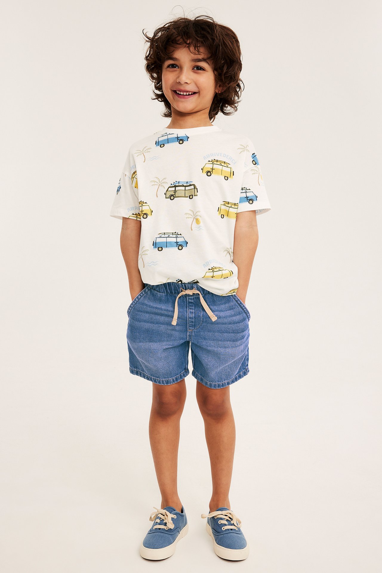 Frontvy av ett barn i mörkblå dra-på-jeansshorts, en vit t-shirt med van-tryck och blå sneakers.