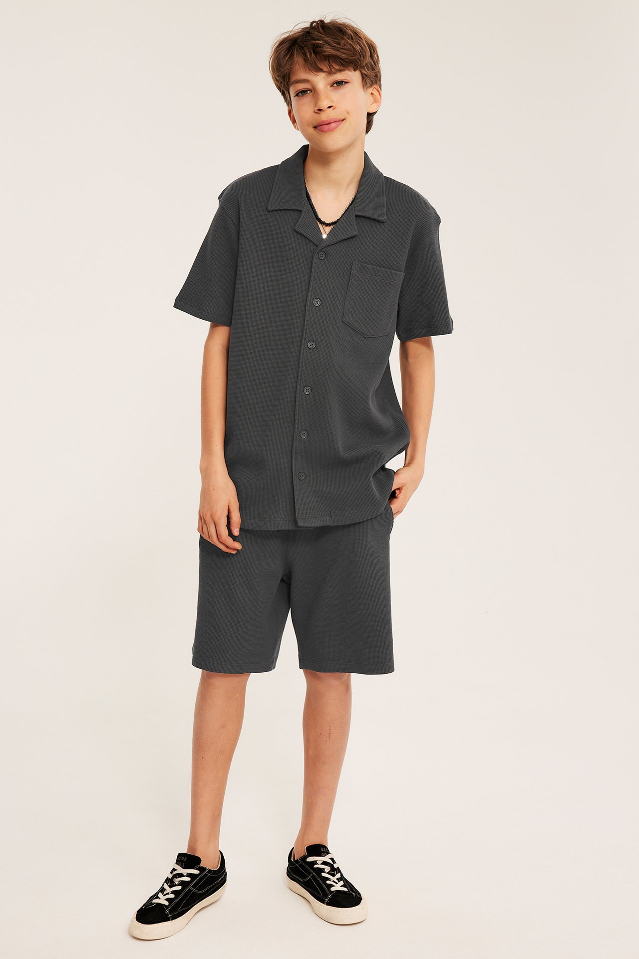 Edestäpäin: Lapsi pukeutuu tummanharmaa vohvelikankaisiin puuvillashortseihin ja yhteensopivaan lyhythihaiseen kauluspaitaan.