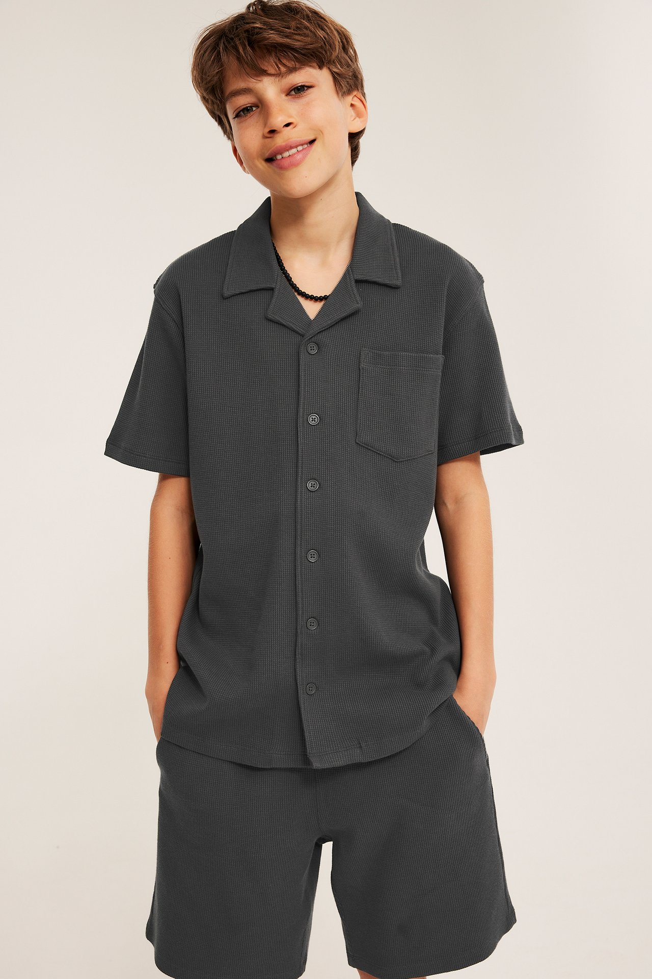 Edestä, lapsi mustissa vohvelineulehortseissa taskuilla ja yhteensopivassa lyhythihaisessa napitussa paidassa.