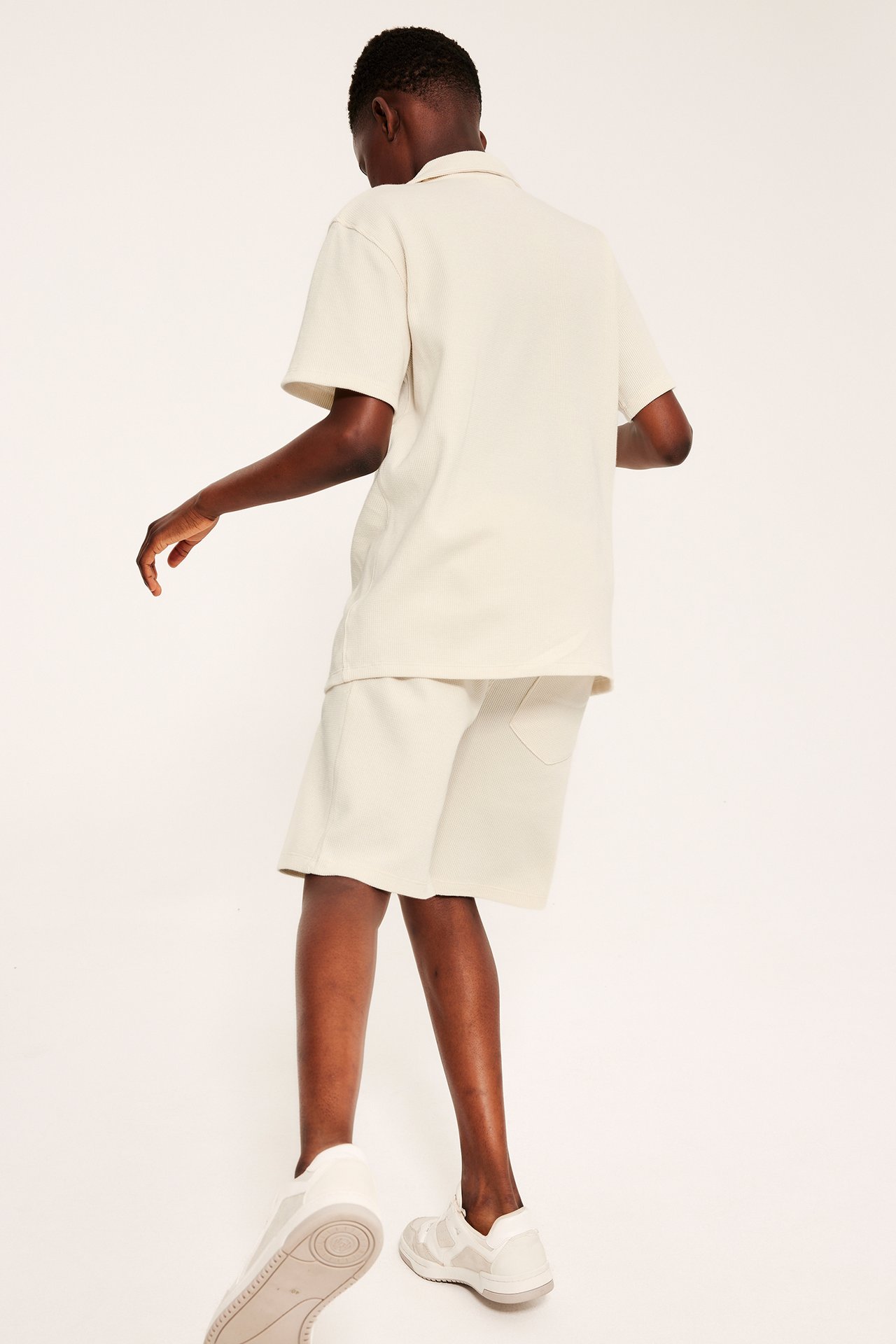 Widok z tyłu modela w jasnobeżowych szortach z bawełny waflowej, pasującej koszulce polo i jasnych sneakersach.