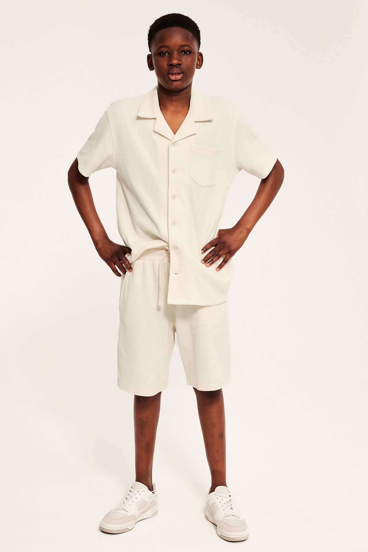 Edestäpäin lapsi vaaleissa vohvelineulotuissa shortseissa, yhteensopivassa paidassa ja valkoisissa tennareissa.