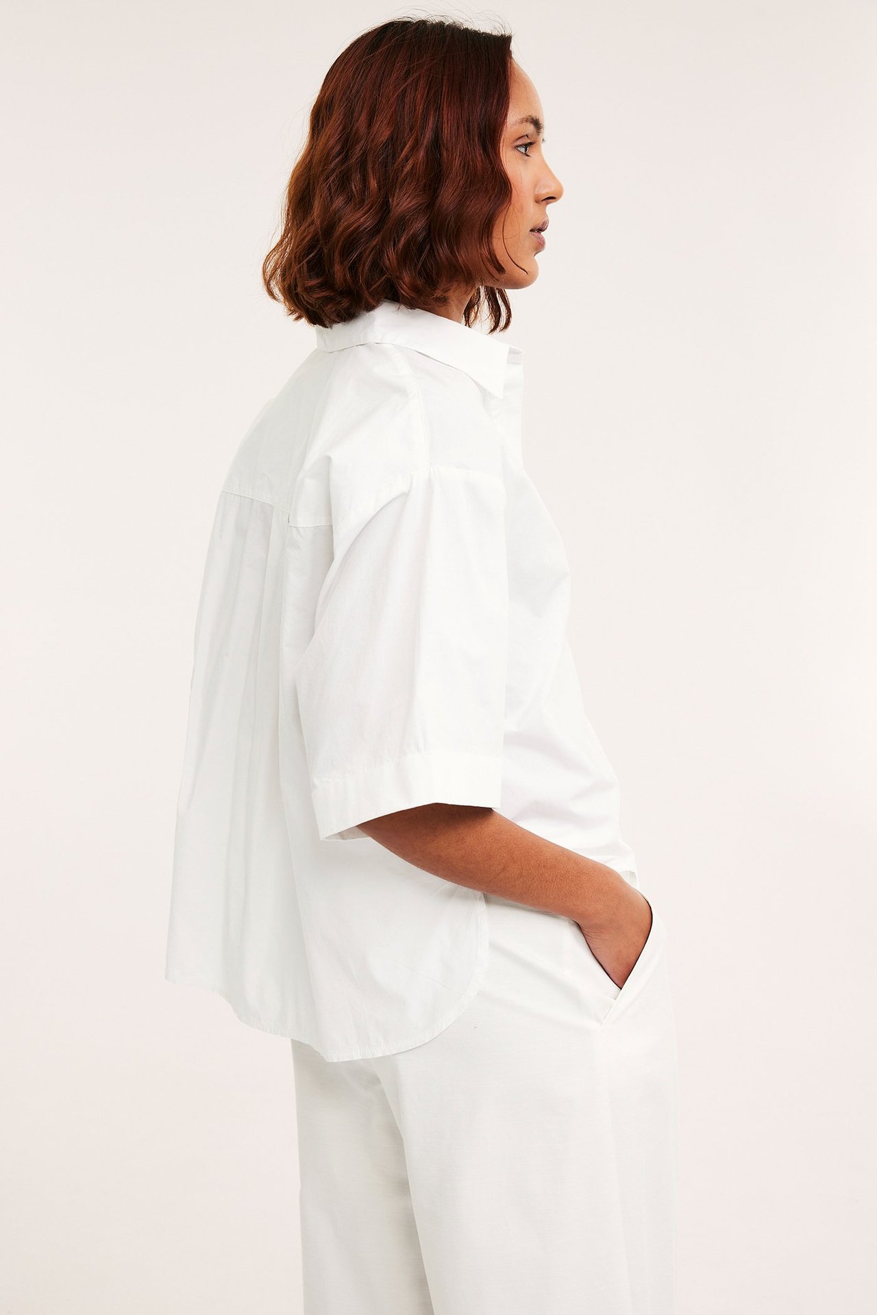 Sidebilde av kvinne i løs hvit kortermet bomullspoplin skjorte med oppbrettede ermer og hvit vid bukse.