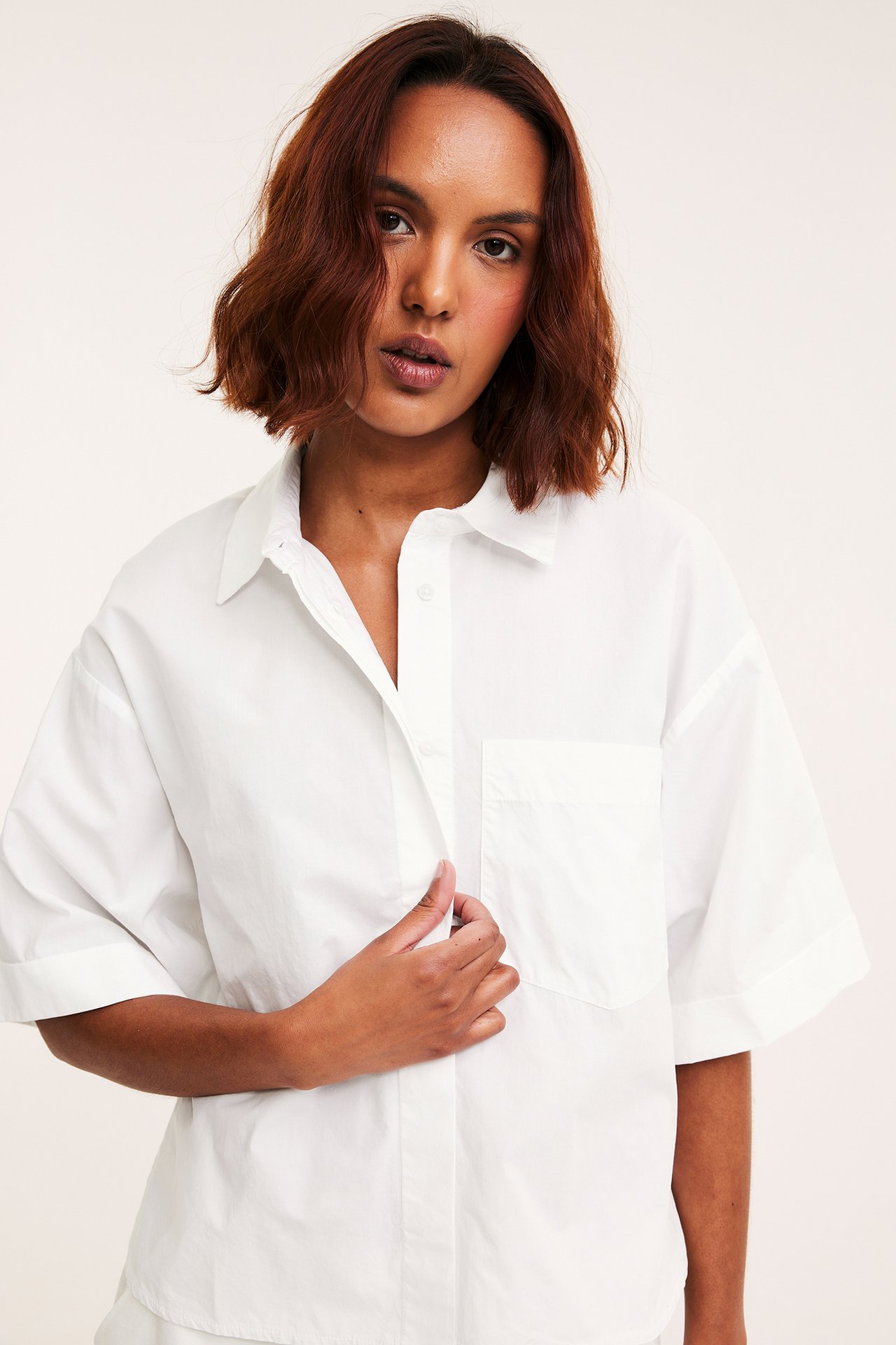 Frontbilde: kvinne i en hvit, løstsittende bomullspoplinskjorte med oppbrettede ermer og brystlomme.