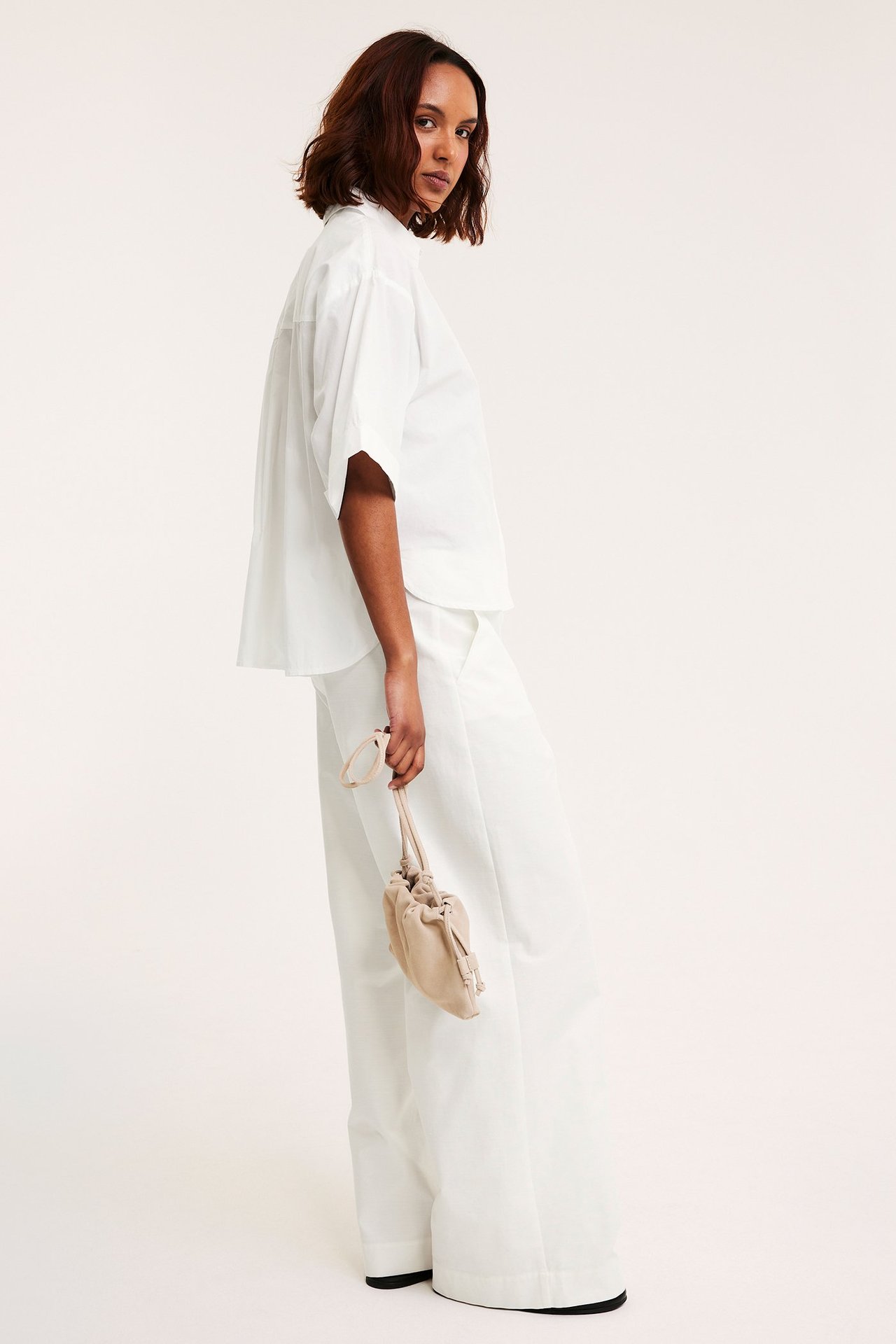 Sidebilde: kvinne i hvit kortermet poplinskjorte, hvite vide bukser, holder beige snøreveske.