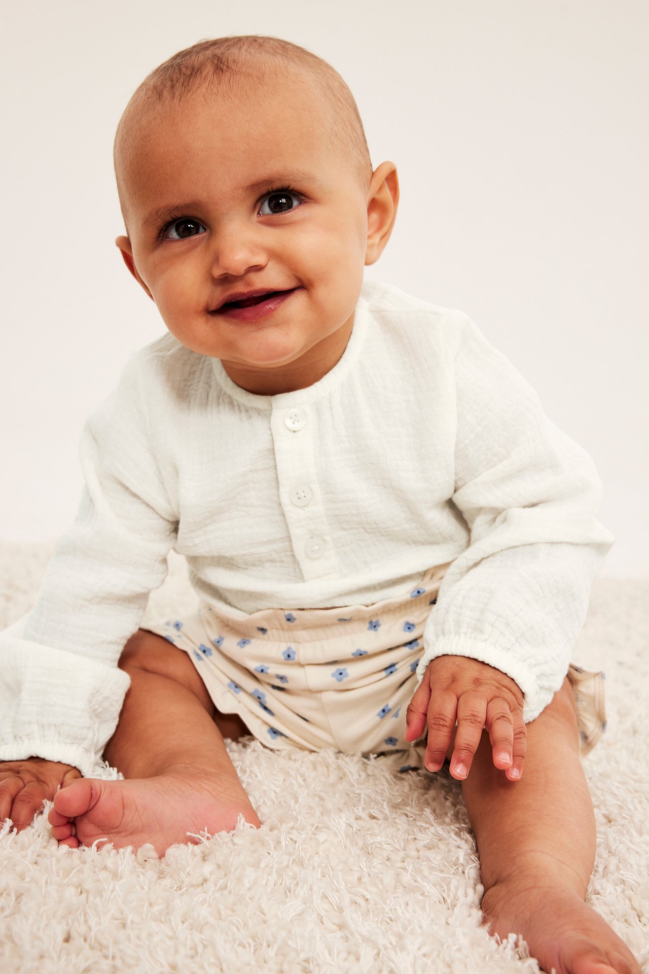 Framsida på leende baby i offwhite långärmad muslinbody och beiga blommiga shorts.