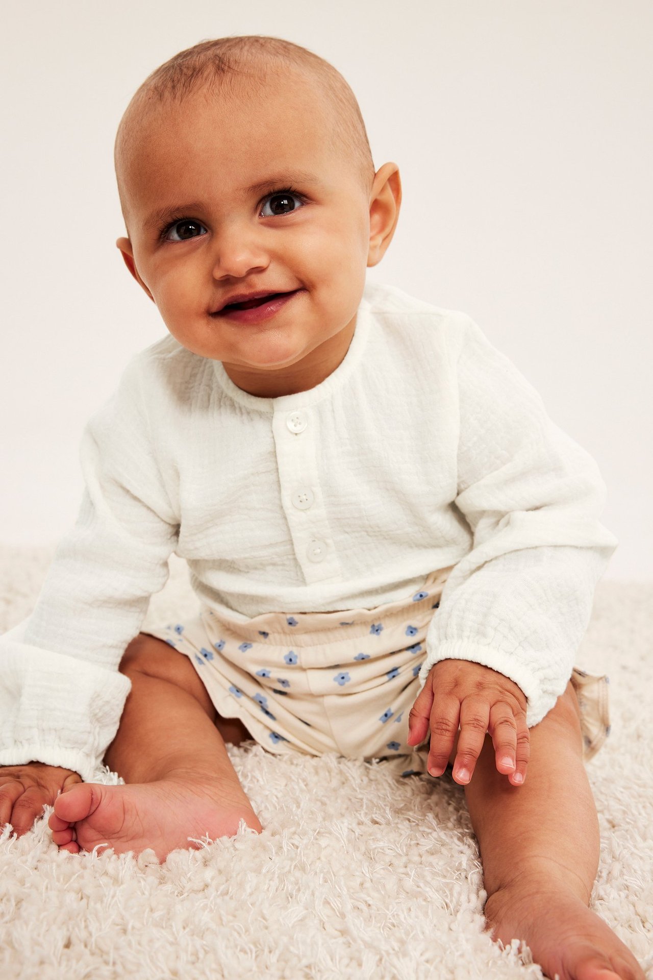 Framsida på leende baby i offwhite långärmad muslinbody och beiga blommiga shorts.